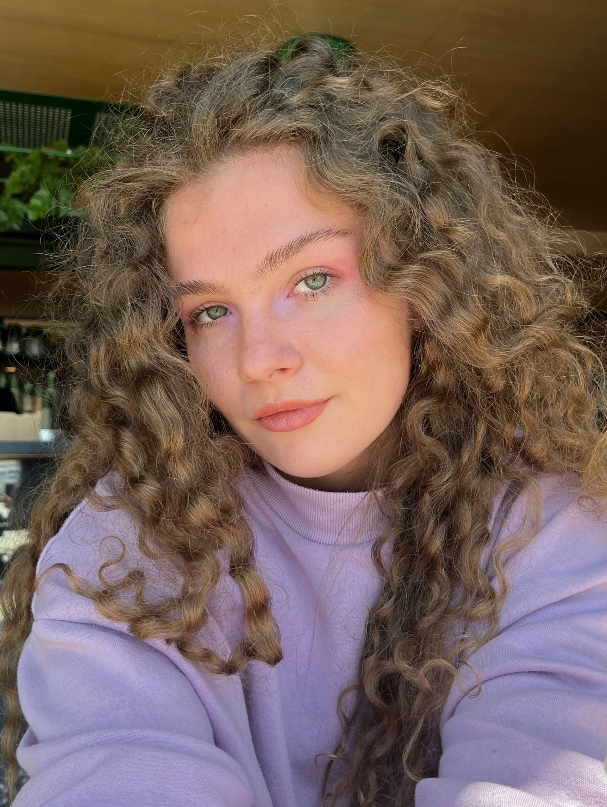 A young woman with curly blonde hair and blue eyes, wearing a light purple shirt, taking a selfie indoors with natural light.