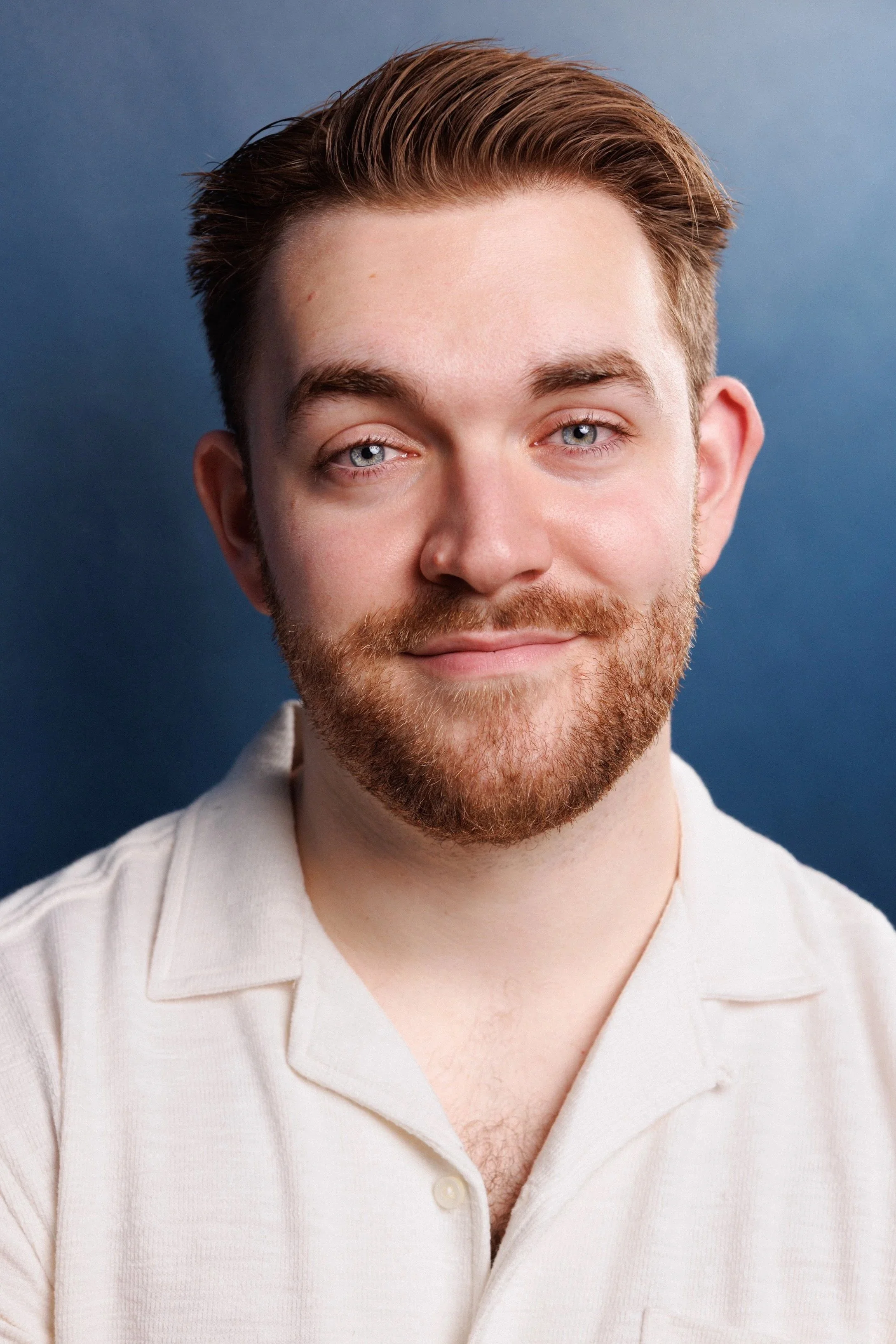 A young man with light skin, brown hair, and a beard, wearing a white shirt, smiling against a blue background.