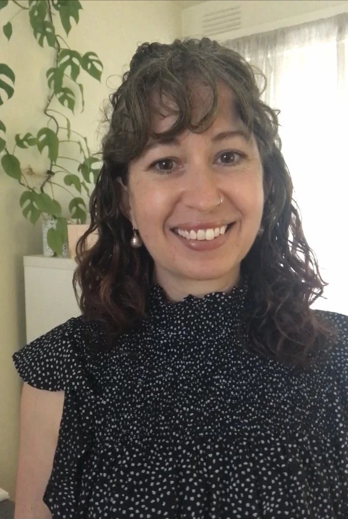 A woman with curly brown hair, smiling, wearing a black dress with white polka dots, earrings, a nose ring, and standing in a room with a plant, a white dresser, and a window with curtains.