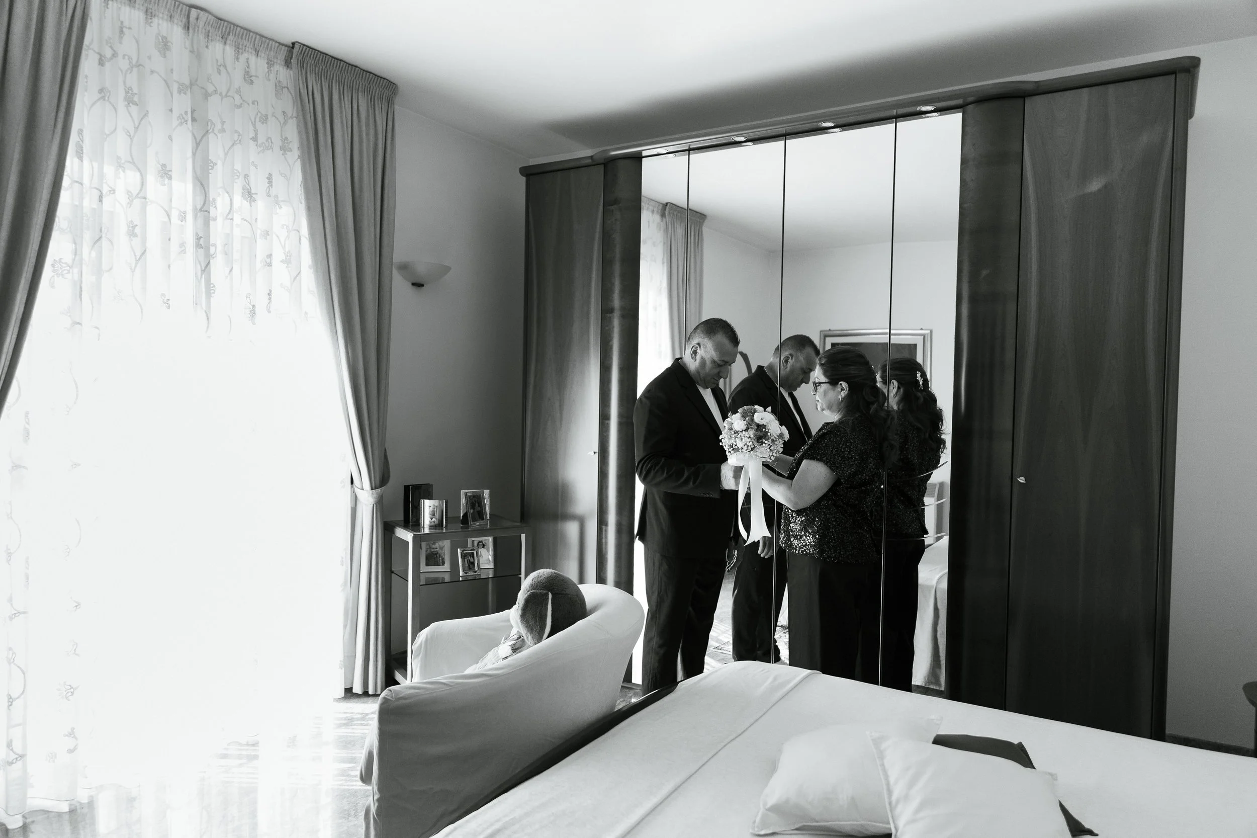 Una coppia si veste con l'aiuto di una donna davanti a uno specchio a parete, mentre una persona anziana è seduta su una poltrona in una stanza luminosa con tende e fotografie sul comodino.