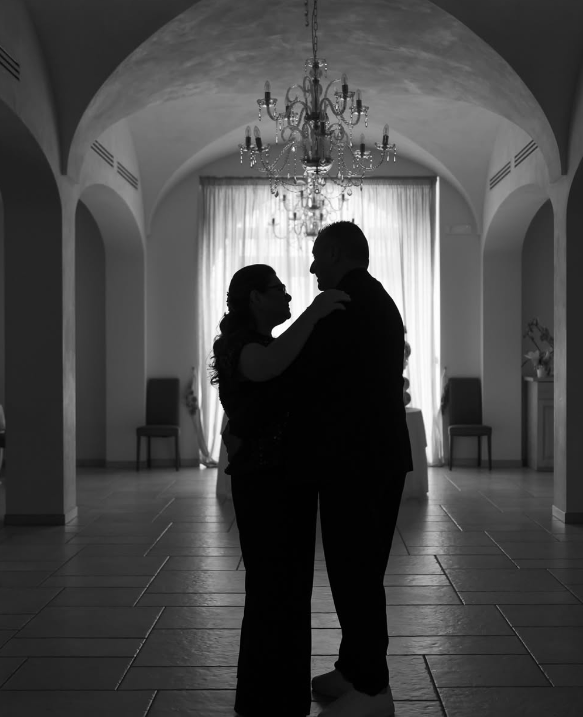 Due persone in silhouette che si abbracciano in una stanza con un chandelier e tende trasparenti sullo sfondo.