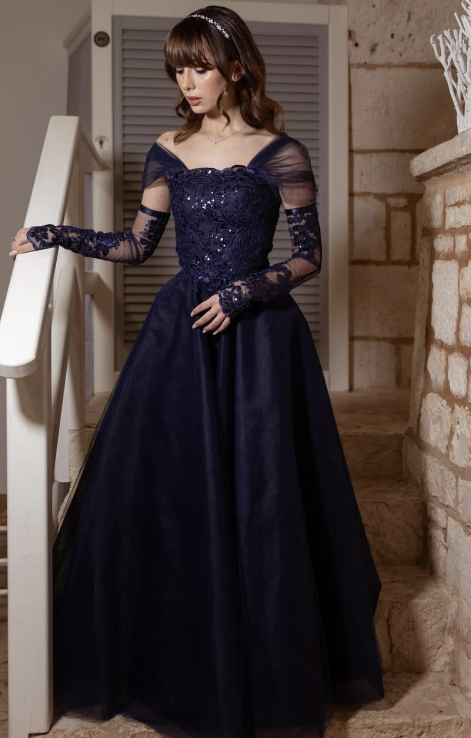 Una donna indossa un elegante abito da sera blu con dettagli in pizzo e maniche lunghe in tulle.