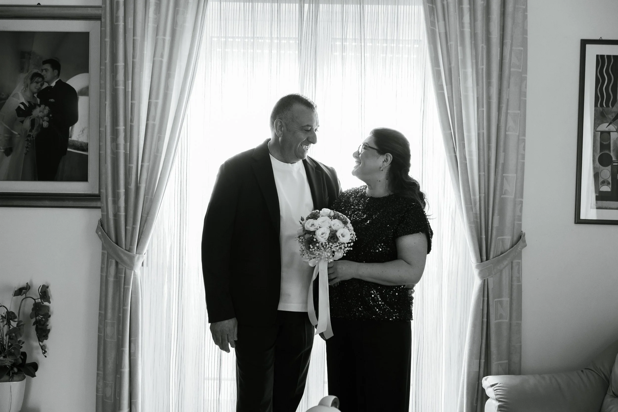 Due persone sorridenti, un uomo e una donna, si guardano e si sorridono mentre la donna tiene un bouquet di fiori. Sono all'interno di una stanza con tende chiare sullo sfondo.