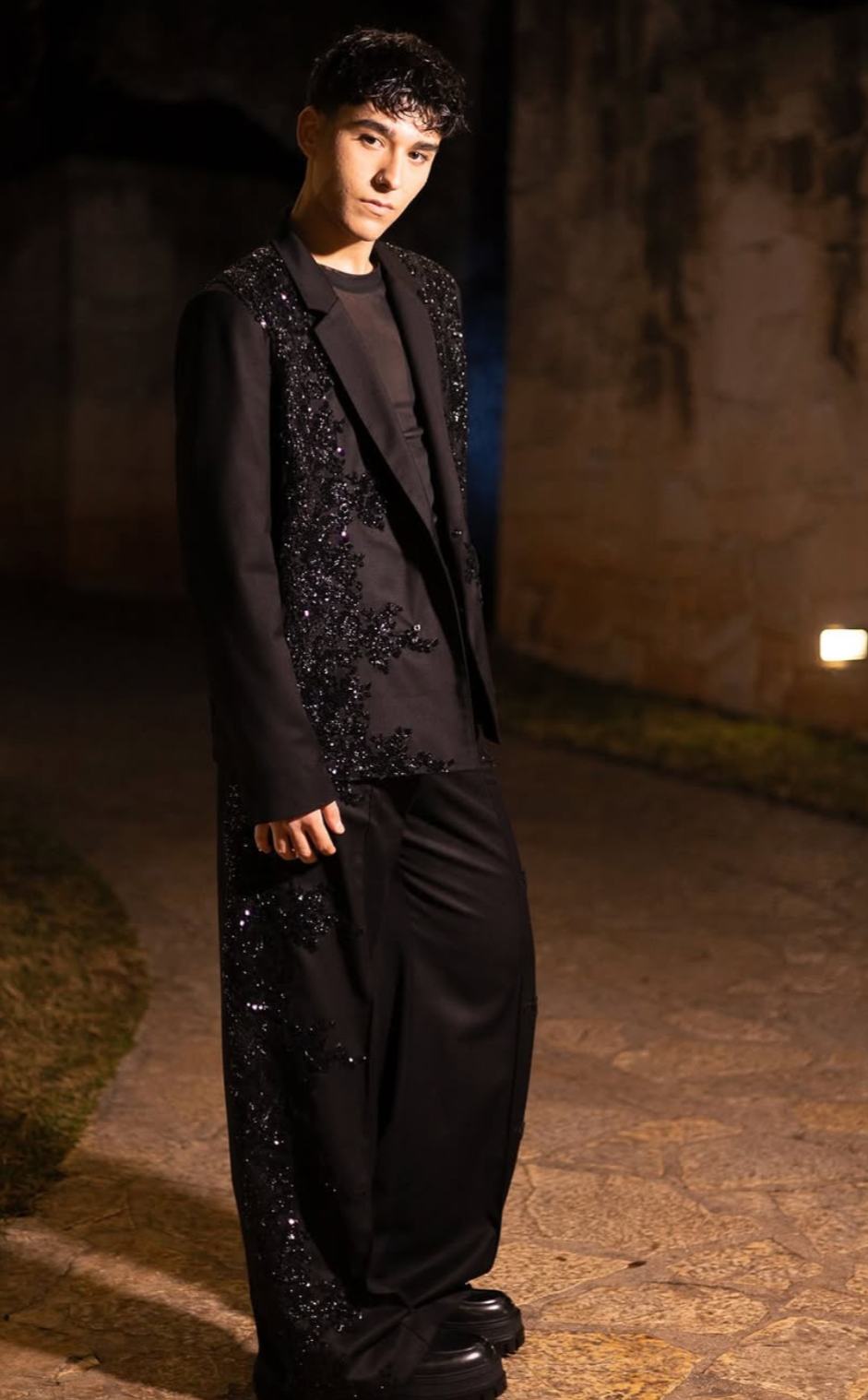 Giovane uomo con capelli ricci neri indossa un abito nero con dettagli di paillettes, in piedi su un percorso di pietra davanti a un muro di pietra, illuminato da luci calde in un ambiente notturno.