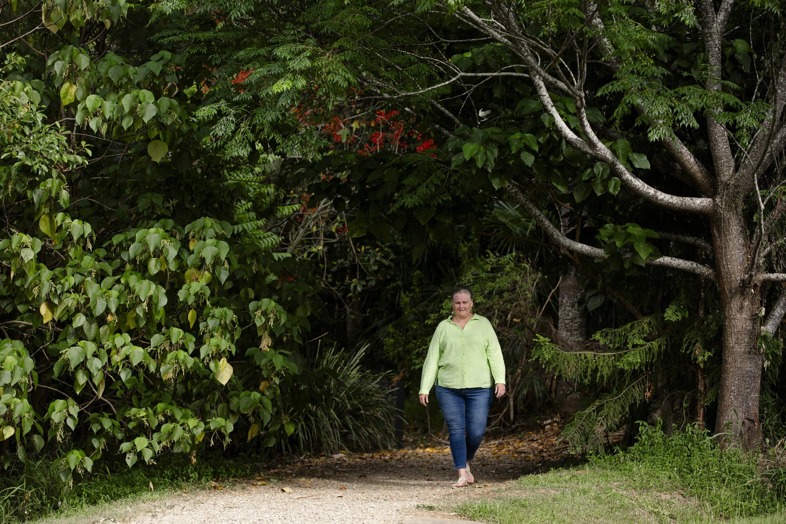 A woman in a light green shirt and blue jeans walking barefoot on a dirt trail through a lush green forest.
