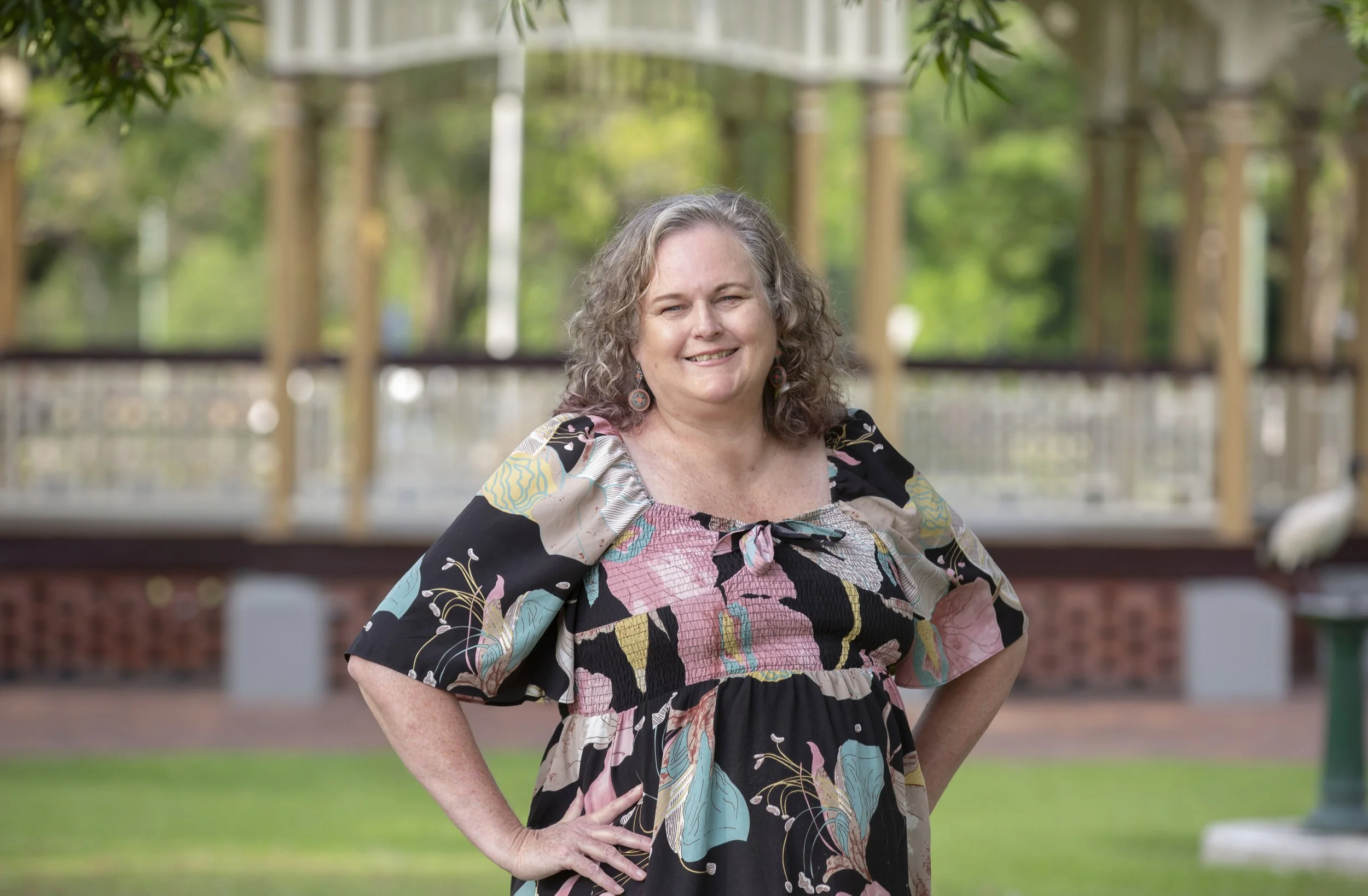 A woman with gray curly hair smiling outdoors, wearing a colorful, patchwork style dress with floral and abstract patterns, in a park setting with green trees and a white bridge in the background.