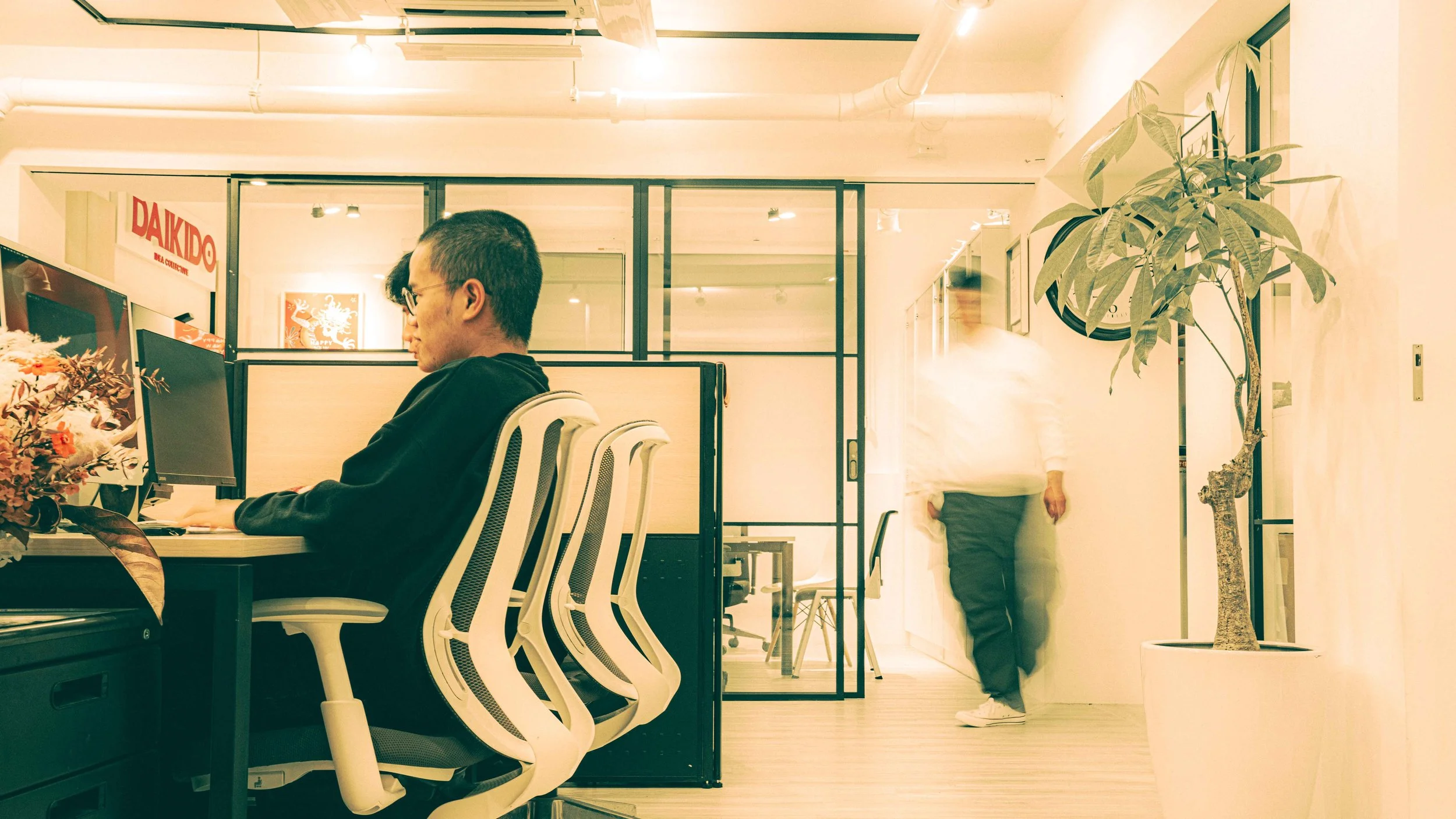 An office space with a person sitting at a desk working on a computer, and another person walking past in the background. There is a large potted plant on the right side and a