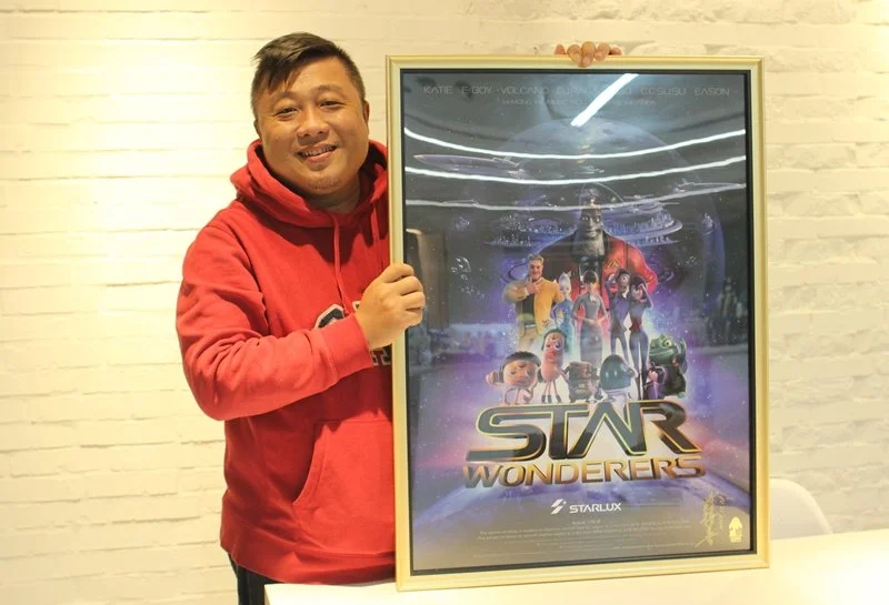 A man in a red hoodie standing next to a framed poster for the animated movie 'Star Wonderers'.