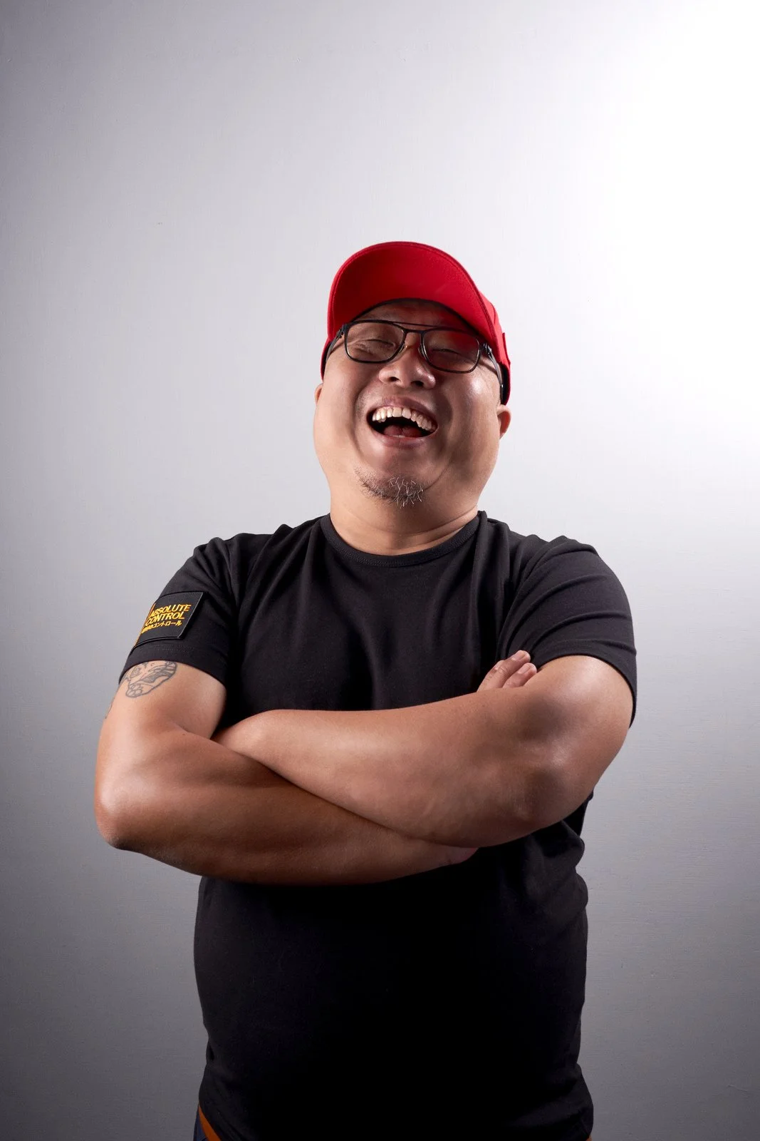 A man with glasses, wearing a red cap and a black t-shirt, smiling with arms crossed against a plain gray background.