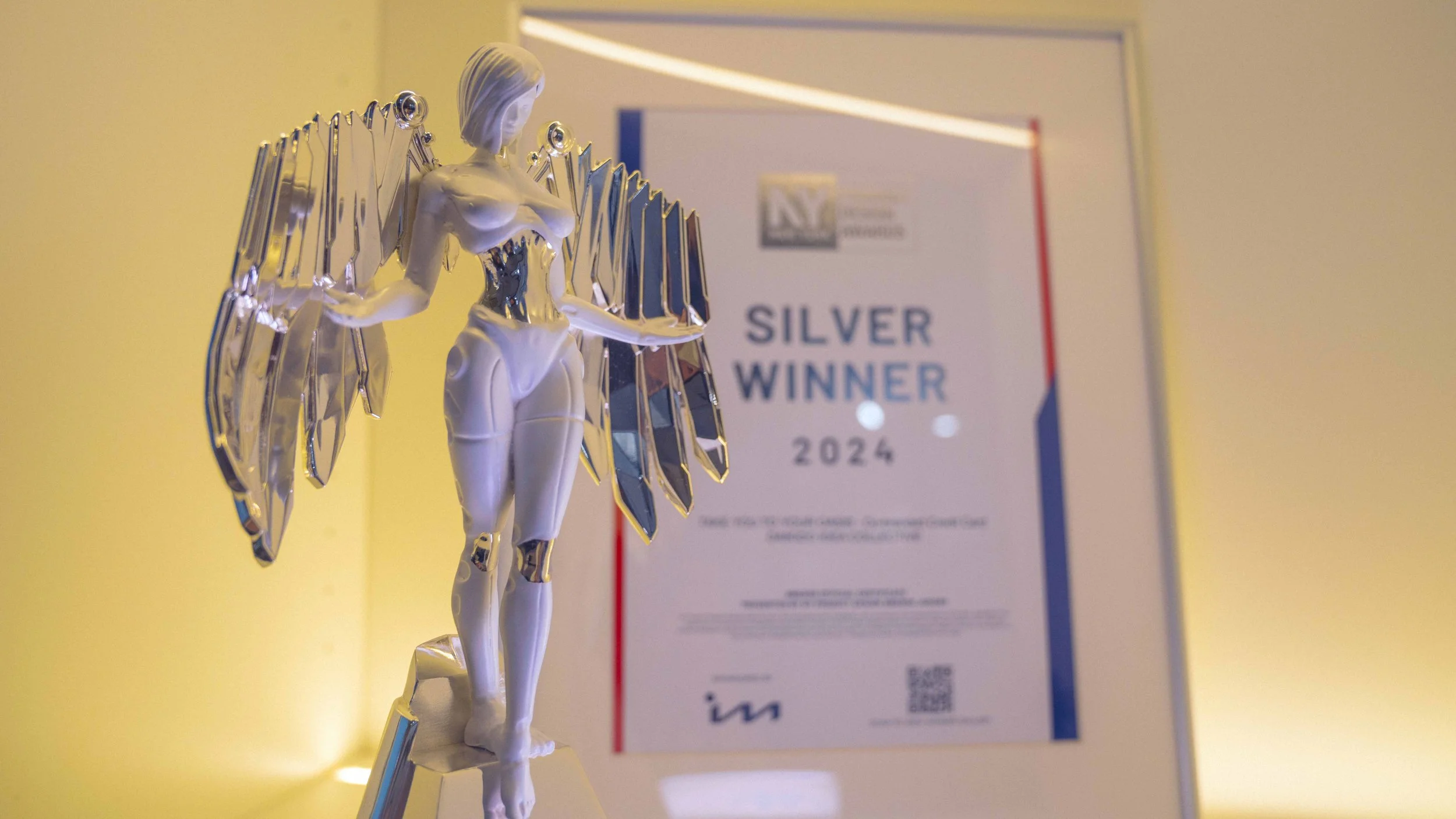 A silver angel trophy with wings made of metallic blades, standing in front of a blurred certificate that reads 'Silver Winner 2024'.