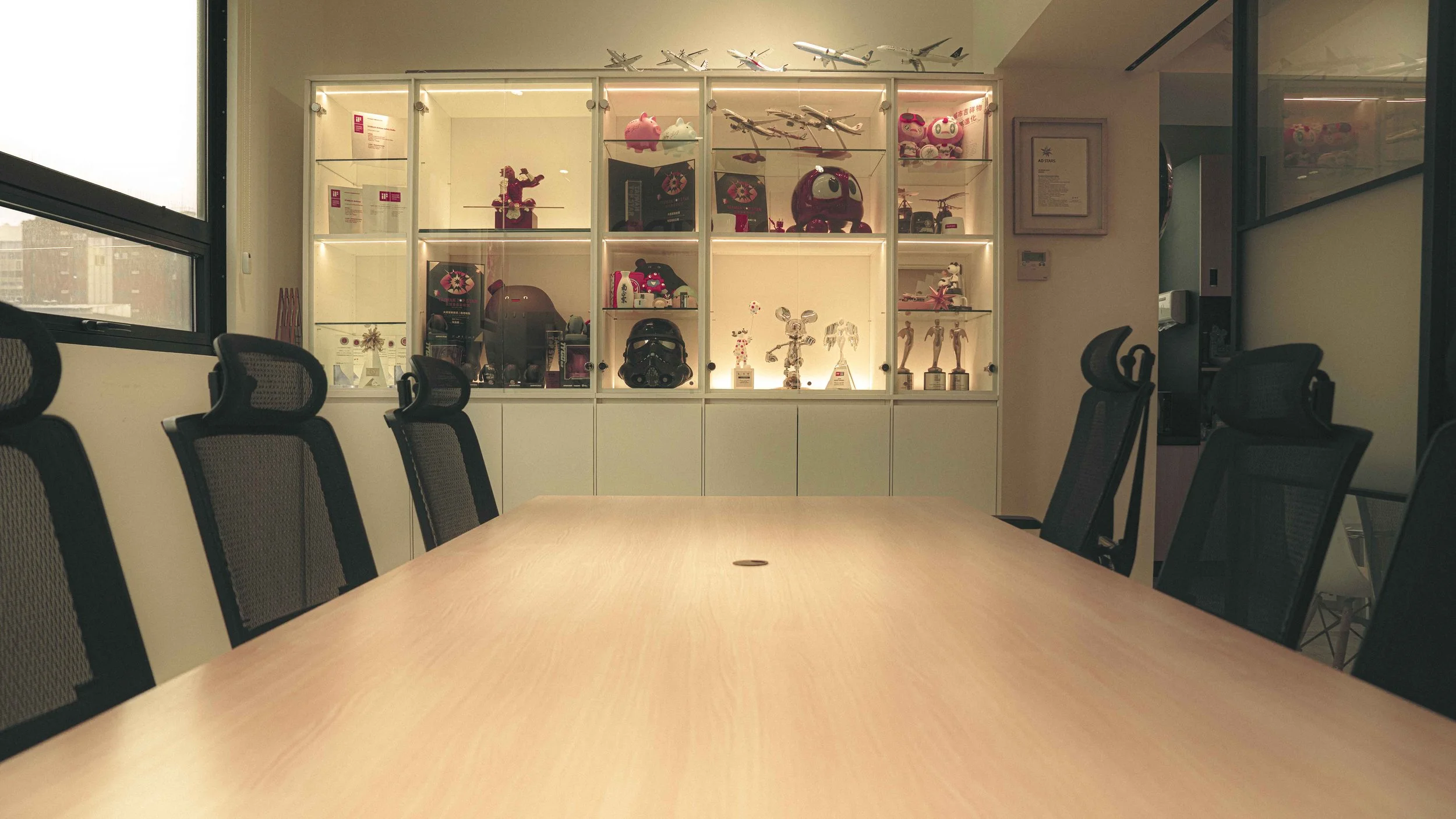 Office conference room with a wooden table and black mesh chairs, and a display cabinet filled with toys and models, illuminated from behind.
