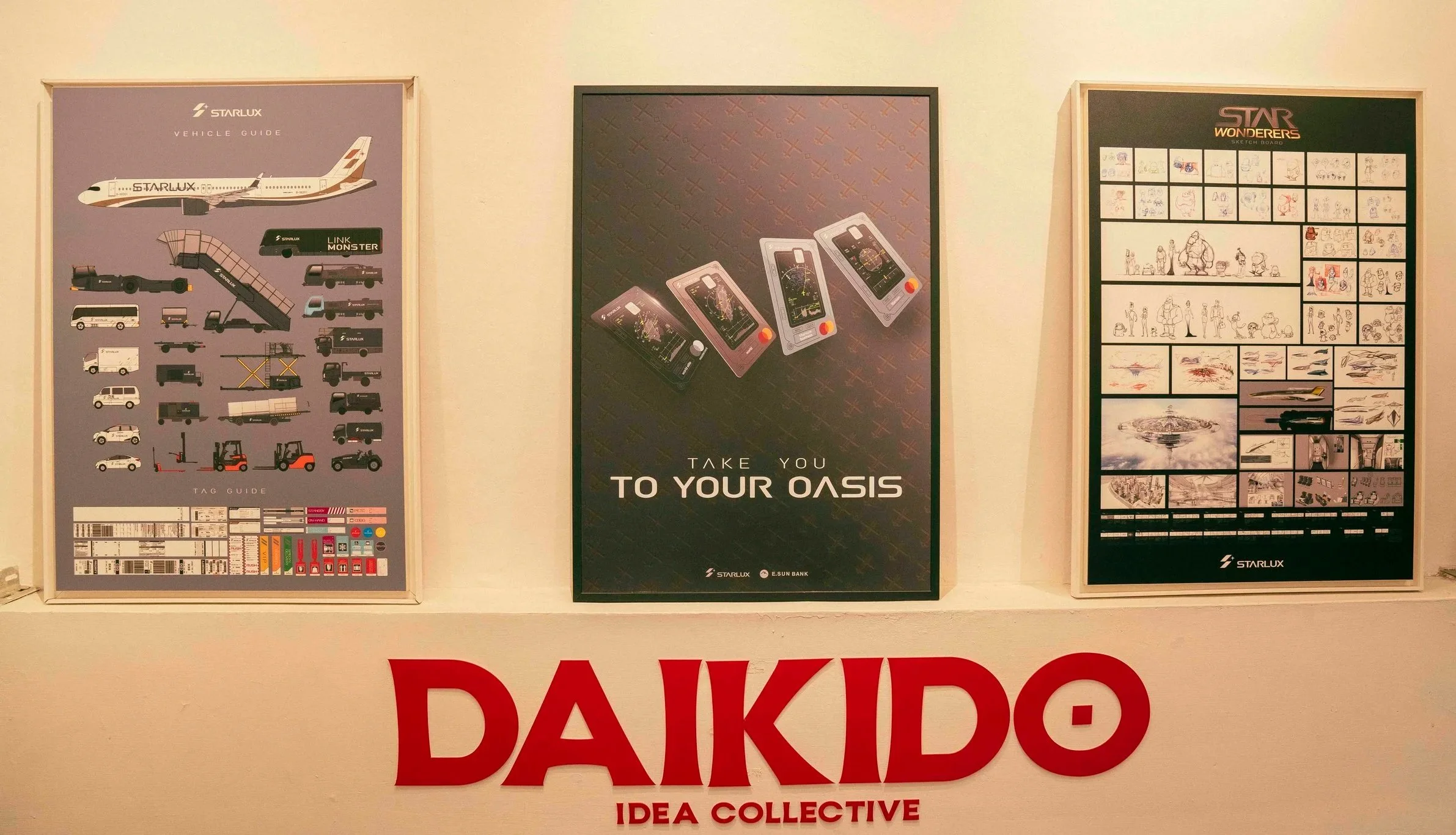 Three posters on a wall behind a red sign that reads 'DAIKIDO IDEA COLLECTIVE.' The first poster shows various aircraft and airport equipment, the second shows three credit cards with the text 'Take You To Your Oasis,' and the third features sketches, drawings, and illustrations of vehicles, spaceships, and people.