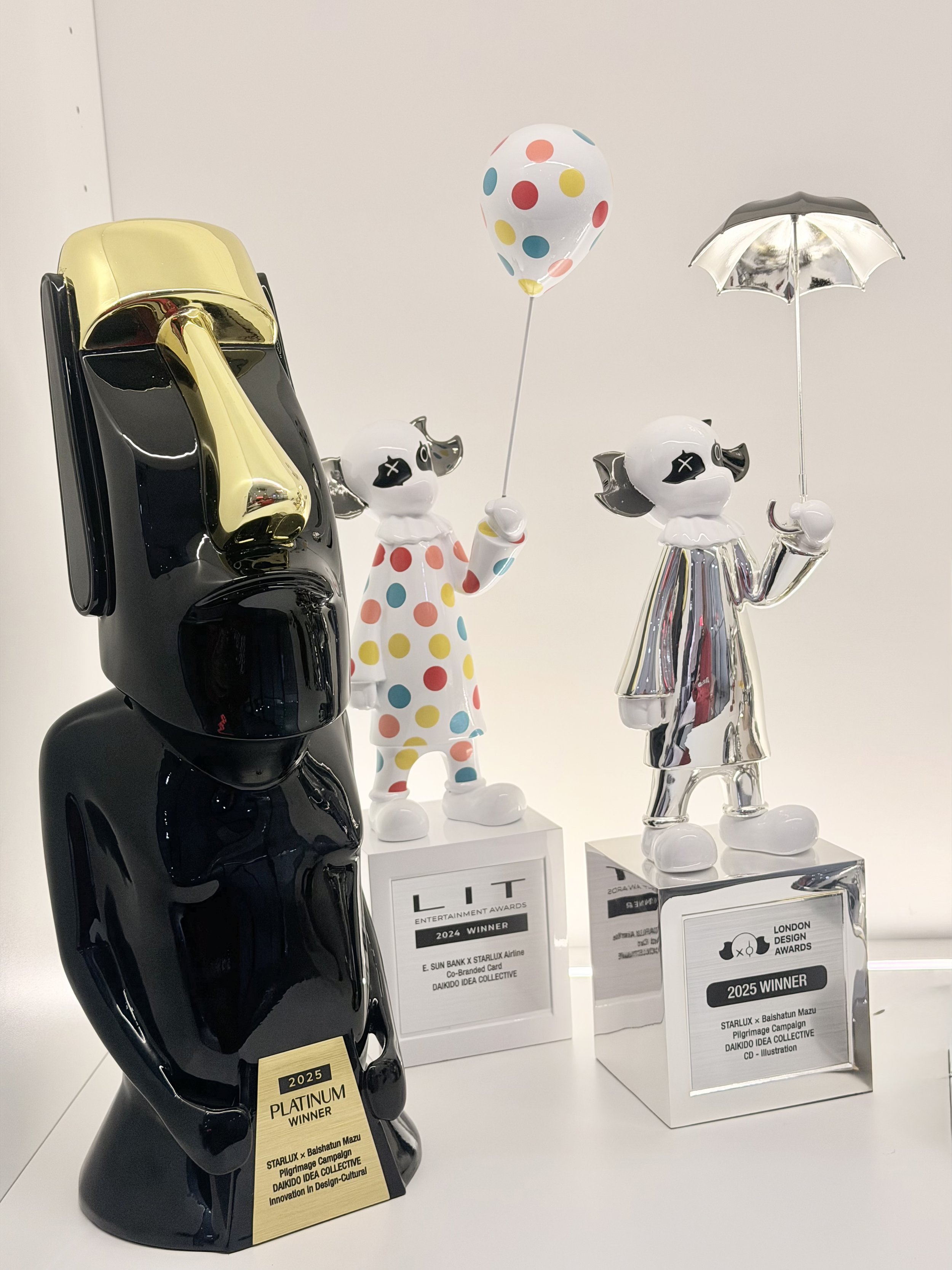 Three decorative award trophies shaped like abstract characters with distinctive faces. The left trophy is black with gold accents, the middle is a white figure with multicolored polka dots holding a polka-dotted balloon, and the right is a silver figure holding a metallic umbrella. Each trophy has a plaque with award details.