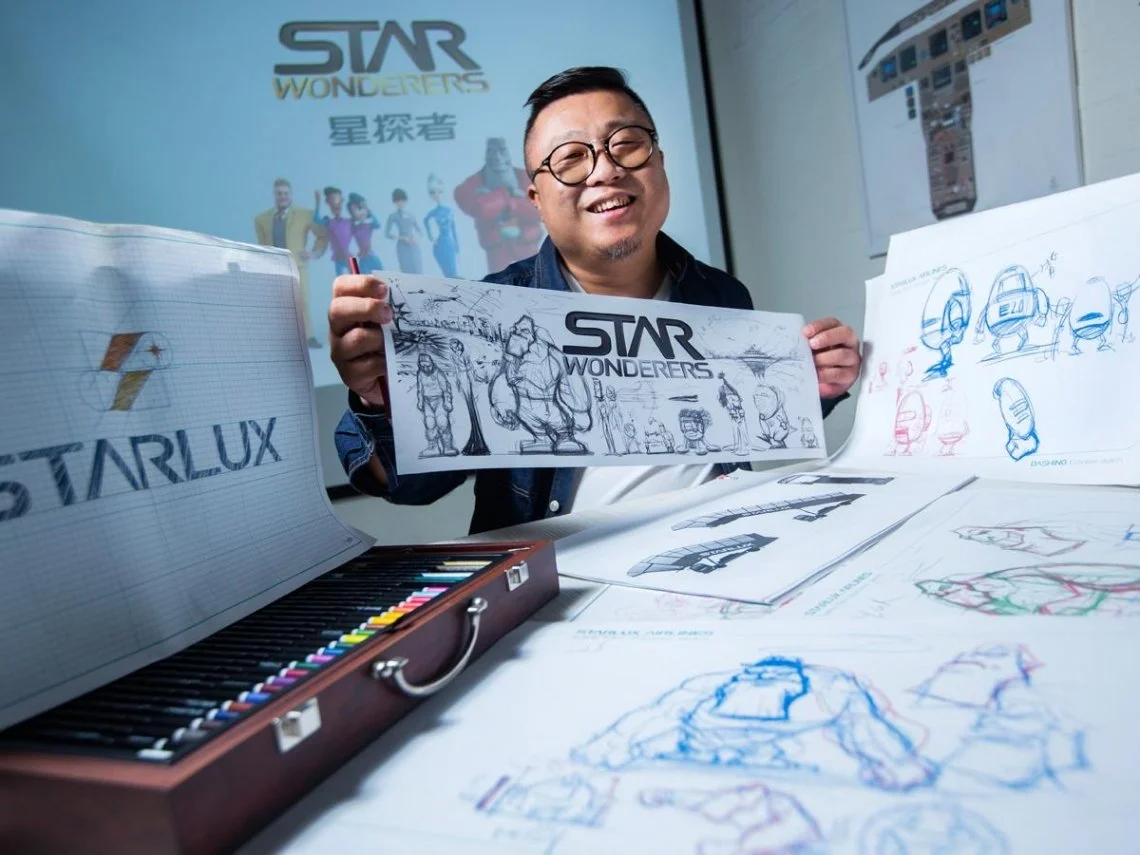 A man with glasses smiling at a table filled with sketches and drawings of Star Wonderers characters and logos, in a creative workspace.