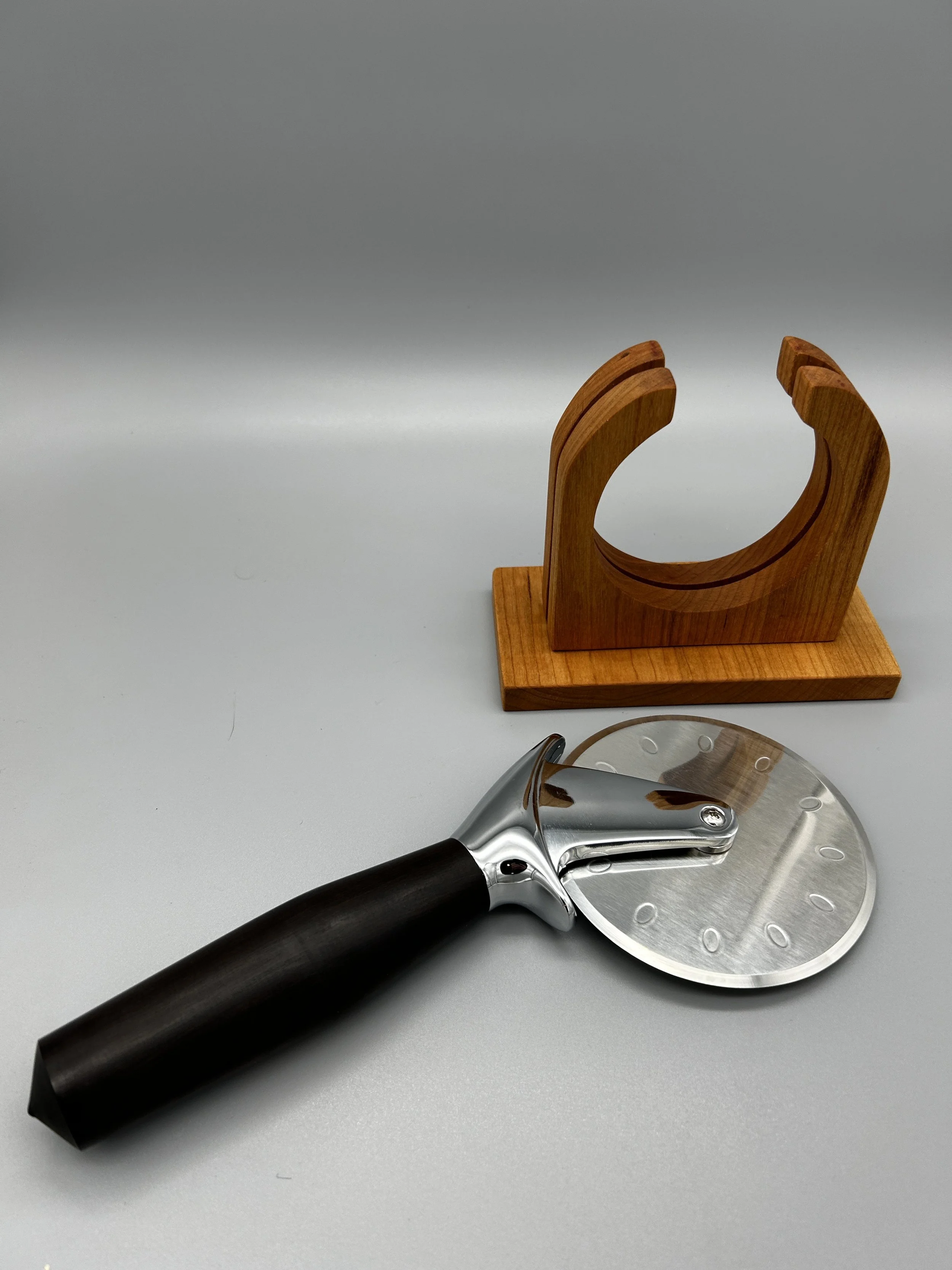 A pizza cutter with a black handle and a wheel with a clock face design sits on a gray surface, with a wooden napkin holder in the background.