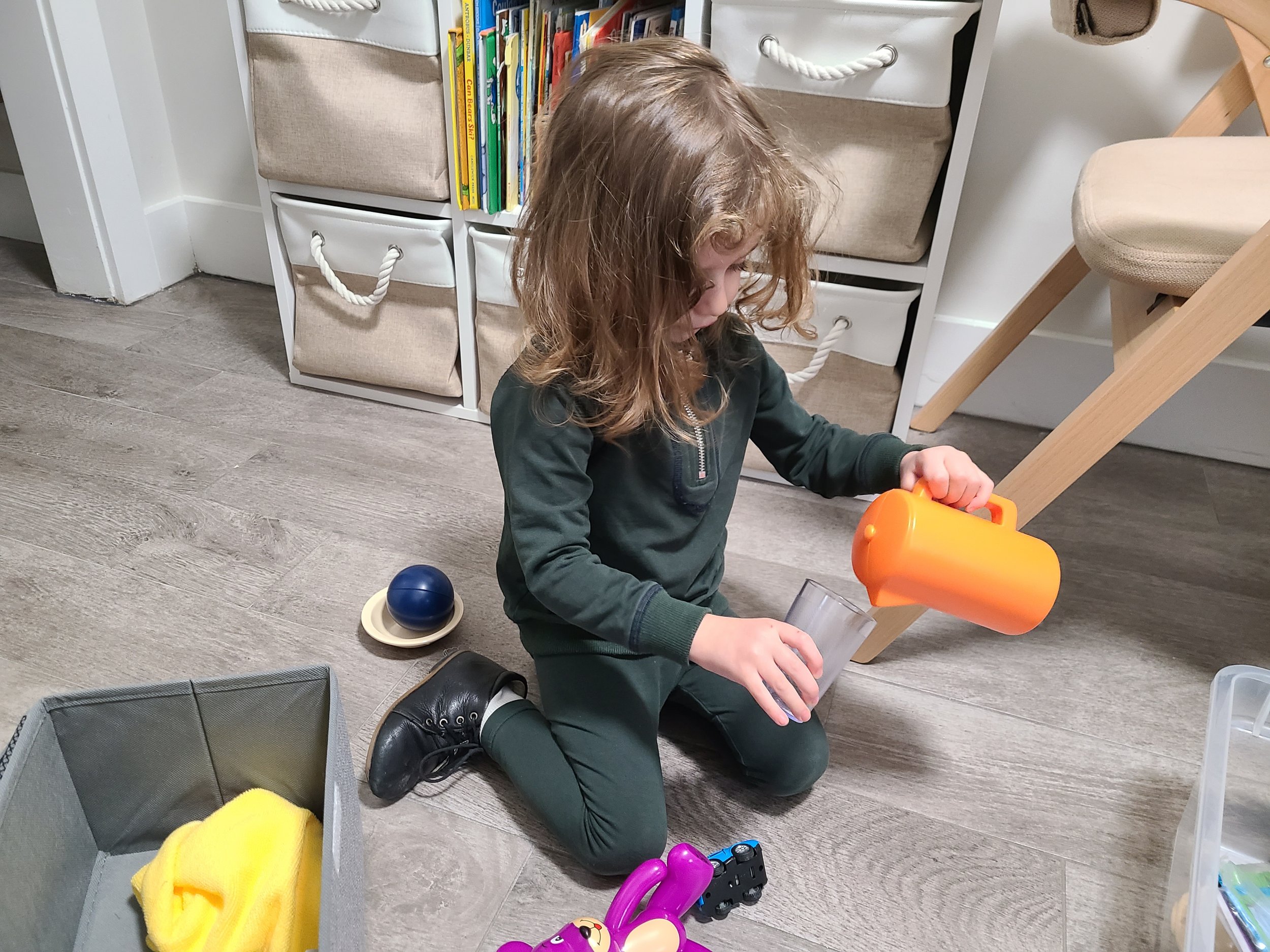 A young girl with curly blonde hair kneeling on a wooden floor, pouring a drink from an orange pitcher into a clear glass. There are various toys and storage bins nearby, with a bookshelf with colorful books in the background.