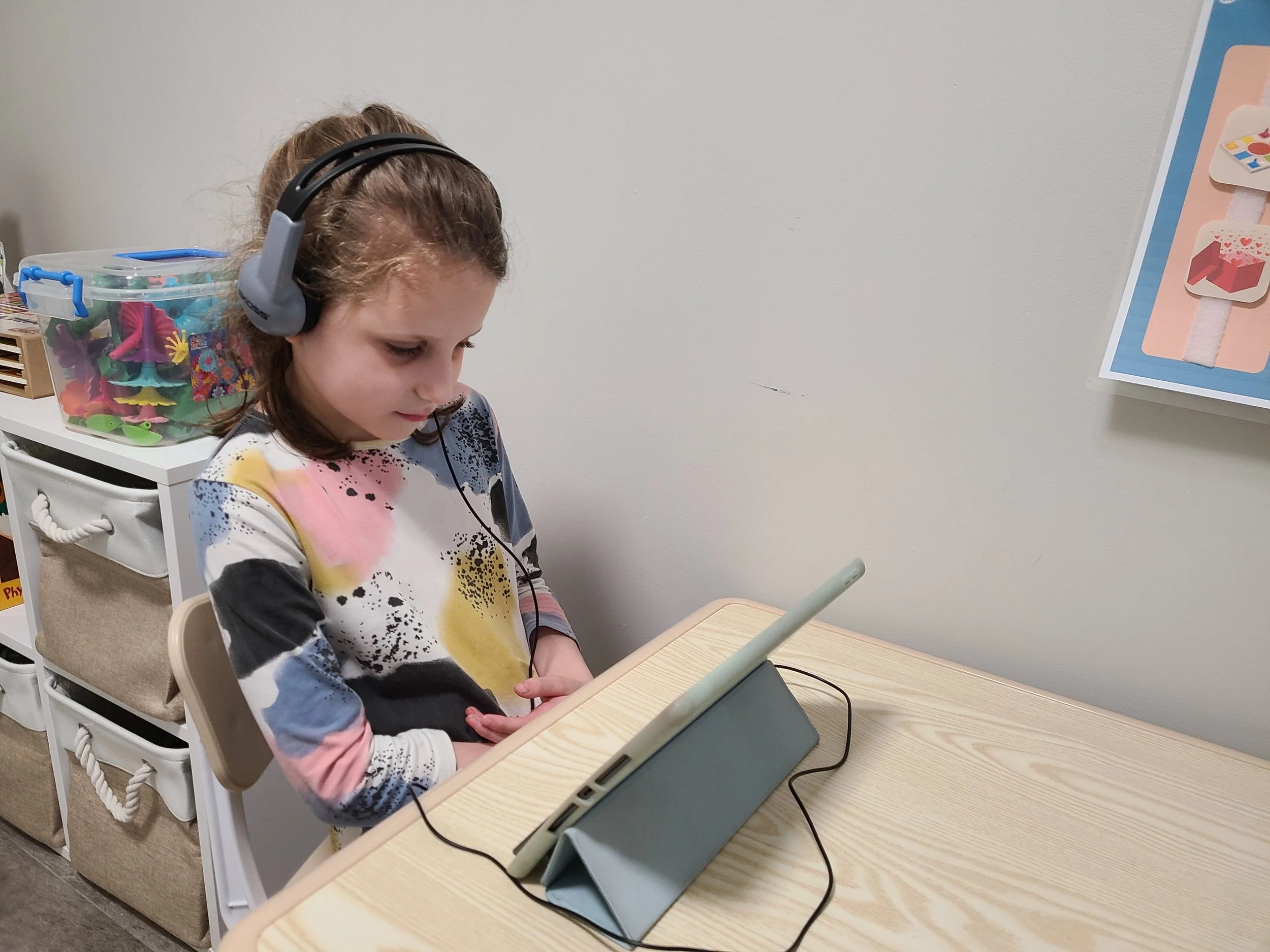 Young girl with headphones looking at a tablet on a light wood desk, with storage bins in the background.