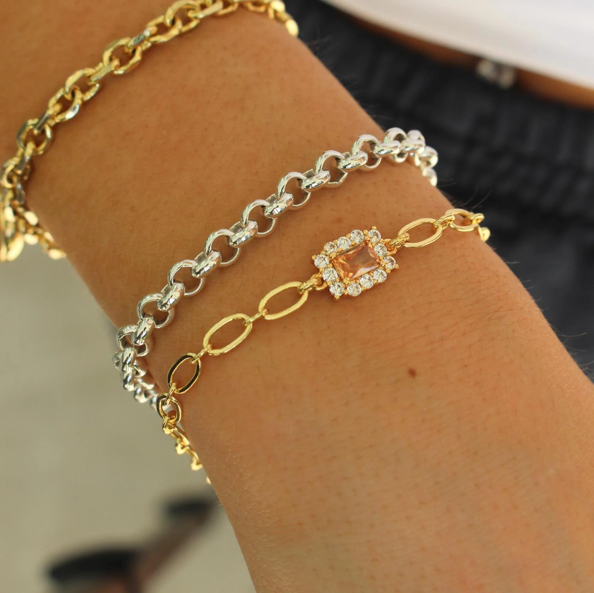 A person's wrist with three layered gold and silver chain bracelets, one featuring a central square gemstone surrounded by small diamonds.