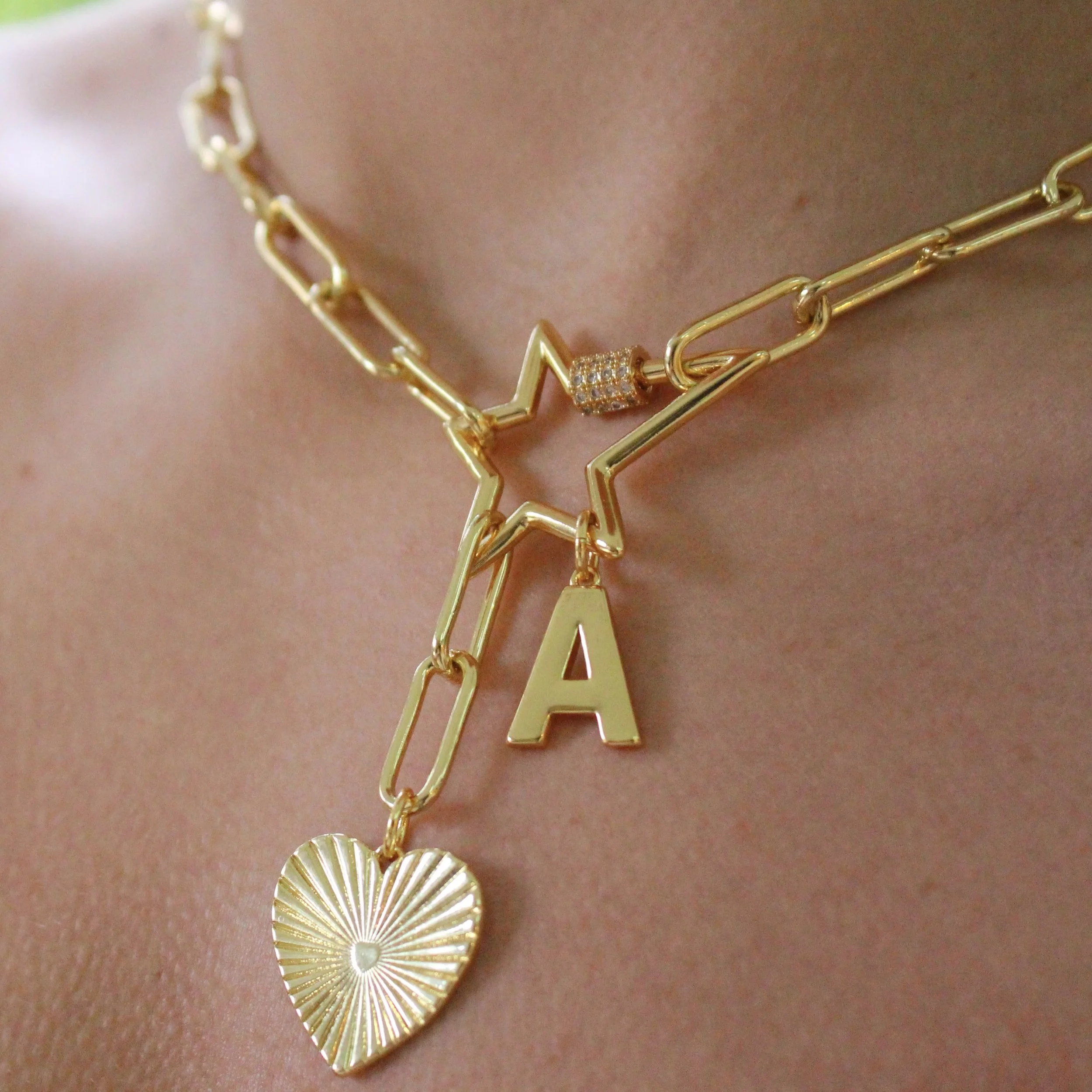 Gold necklace with a star-shaped charm, a letter 'A' charm, a heart-shaped charm, and a small cubic bead, worn around the neck of a person.