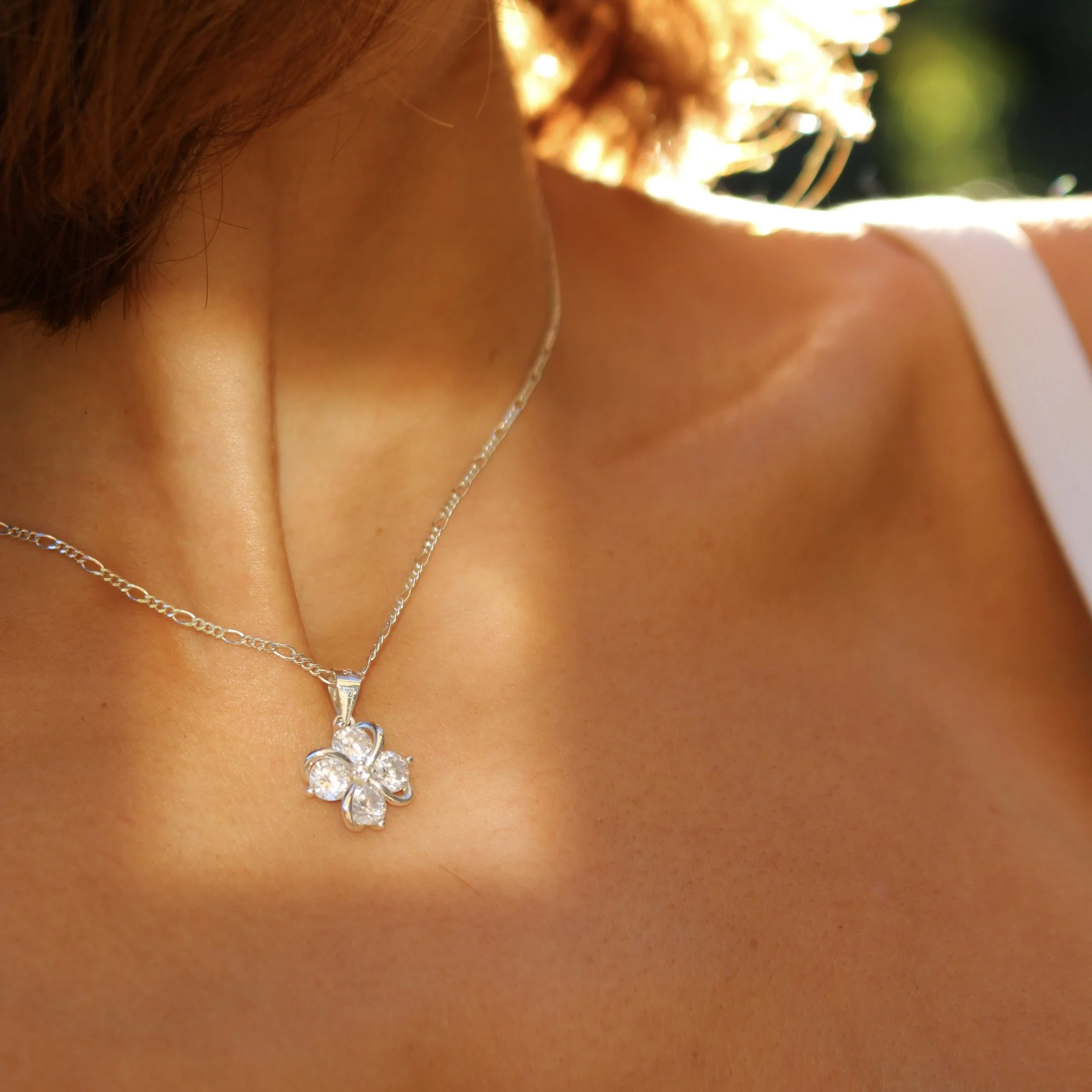 Close-up of a woman's neck and shoulder wearing a silver necklace with a four-leaf clover pendant adorned with clear gemstones, sunlight highlighting her skin.