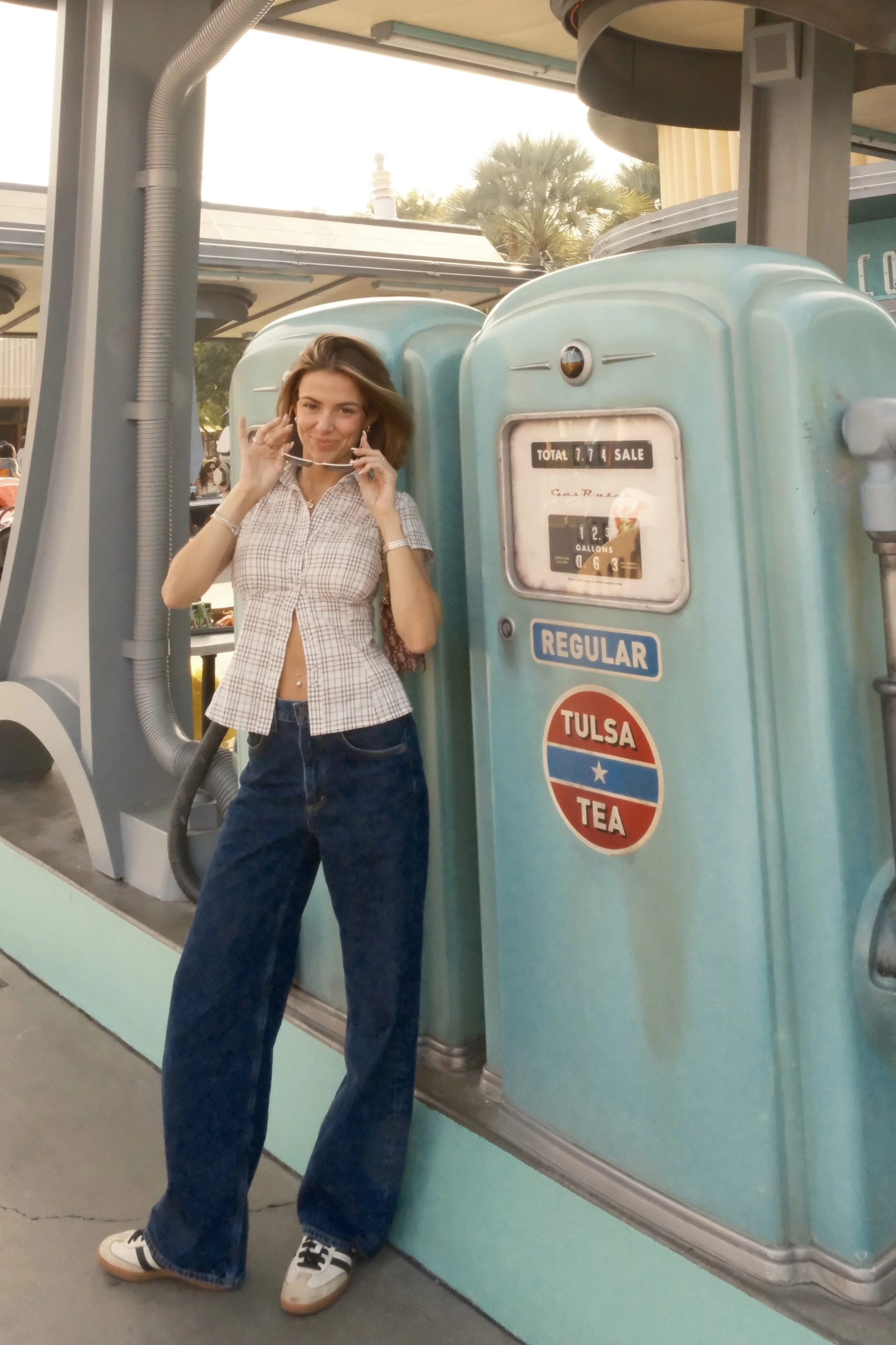 Woman standing beside vintage teal gas pump with 'Tulsa Tea' sticker, adjusting sunglasses, at an outdoor location with palm trees in background.