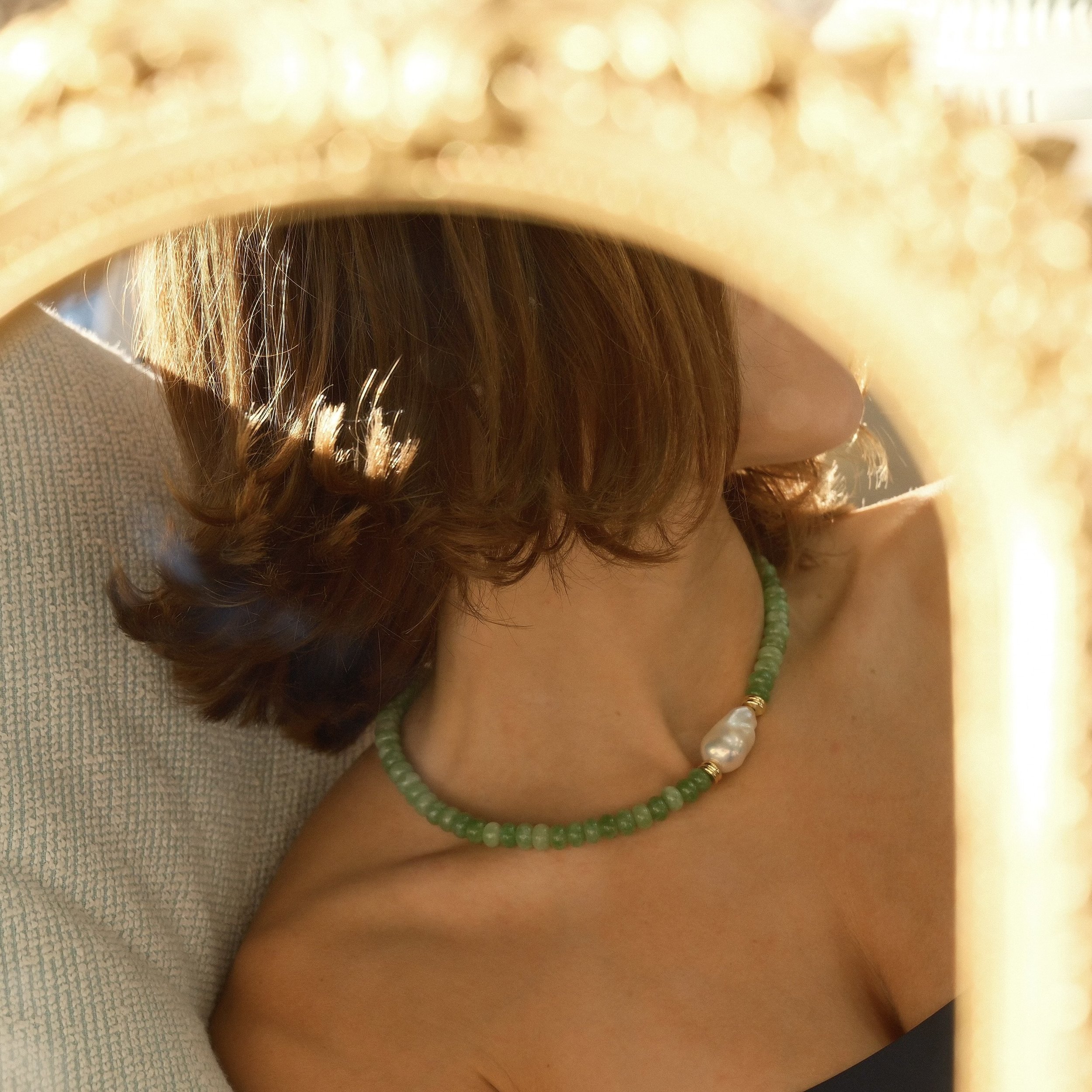 A woman with curly brown hair wearing a green beaded necklace with a large white pearl in the center, sitting on a beige textured chair, seen through a circular, gold-colored mirror frame.