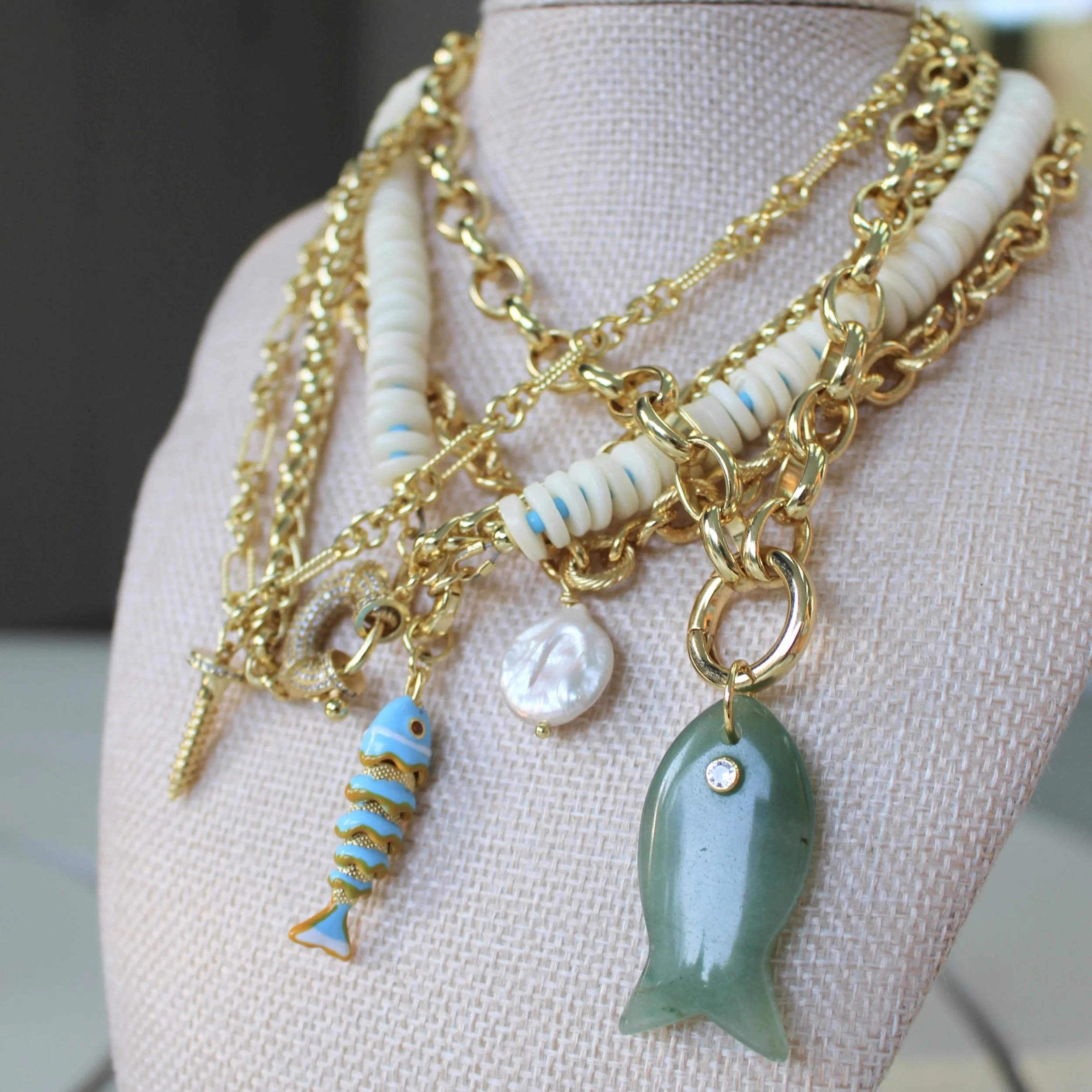 Multiple layered gold chains with pendants, including a fish, a shell, and a green gemstone, displayed on a jewelry bust.