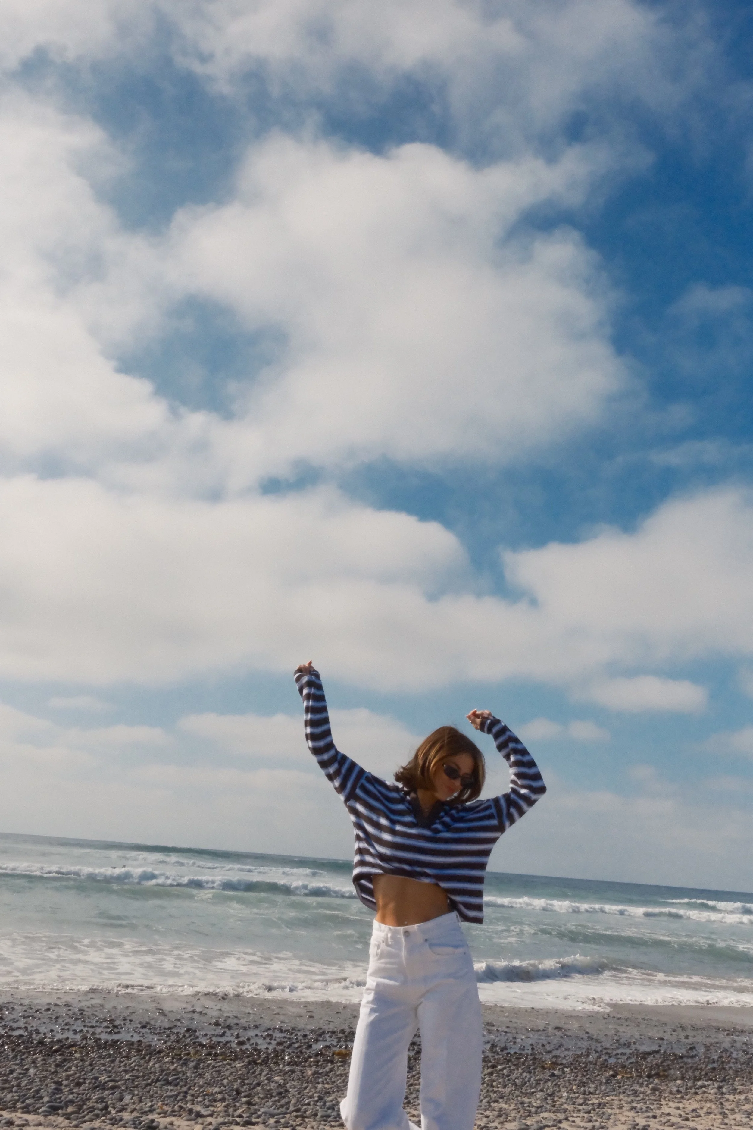 A woman standing on a pebble beach with ocean waves and a cloudy sky in the background. She is wearing sunglasses, a cropped striped long-sleeve shirt, and white pants, with her arms raised and flexed.