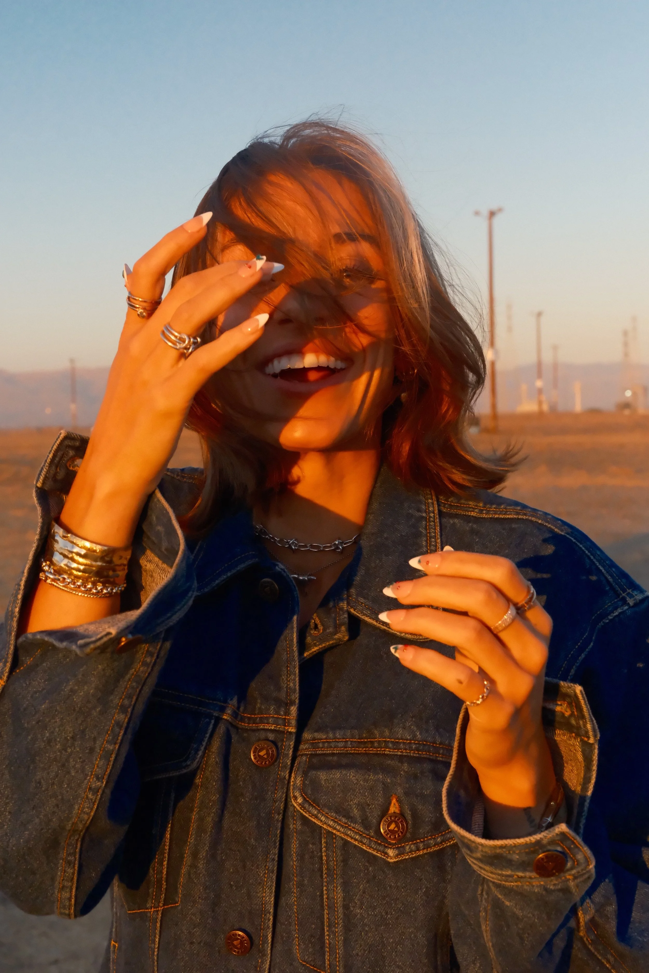 A woman with shoulder-length hair smiling and laughing outdoors at sunset, wearing a denim jacket and multiple rings and bracelets.