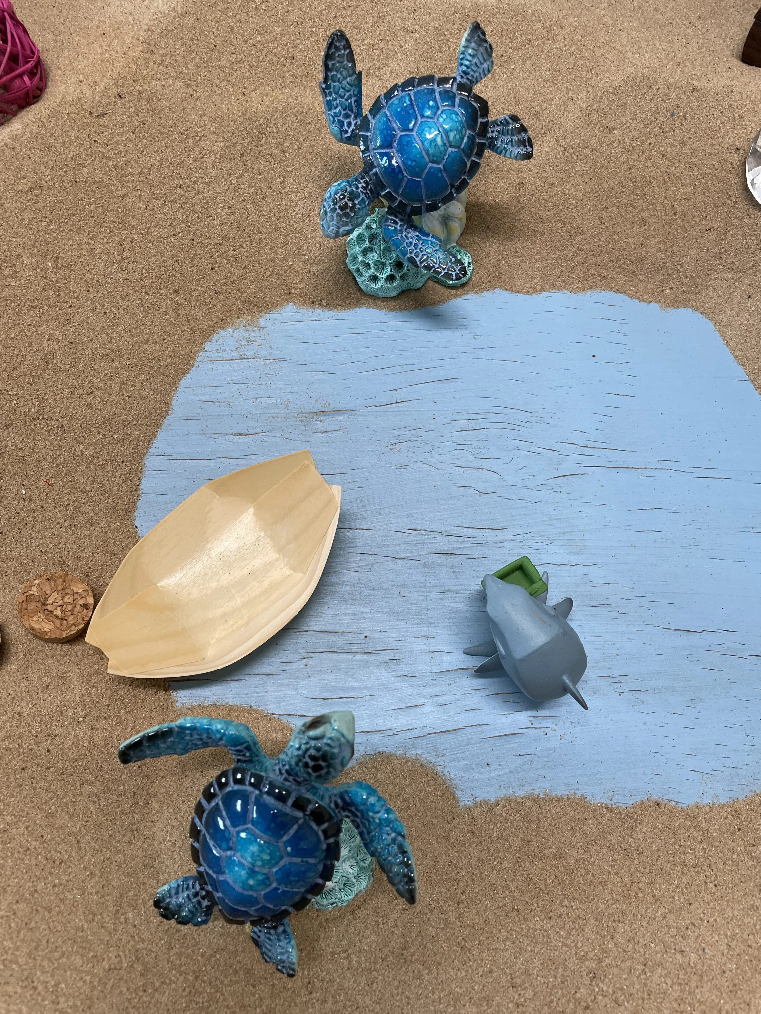 Toy sea creatures and accessories arranged on a sandy surface with a painted blue area. Two blue plastic turtles, a gray dolphin, a beige shell, and a cork ball. sandtray therapy