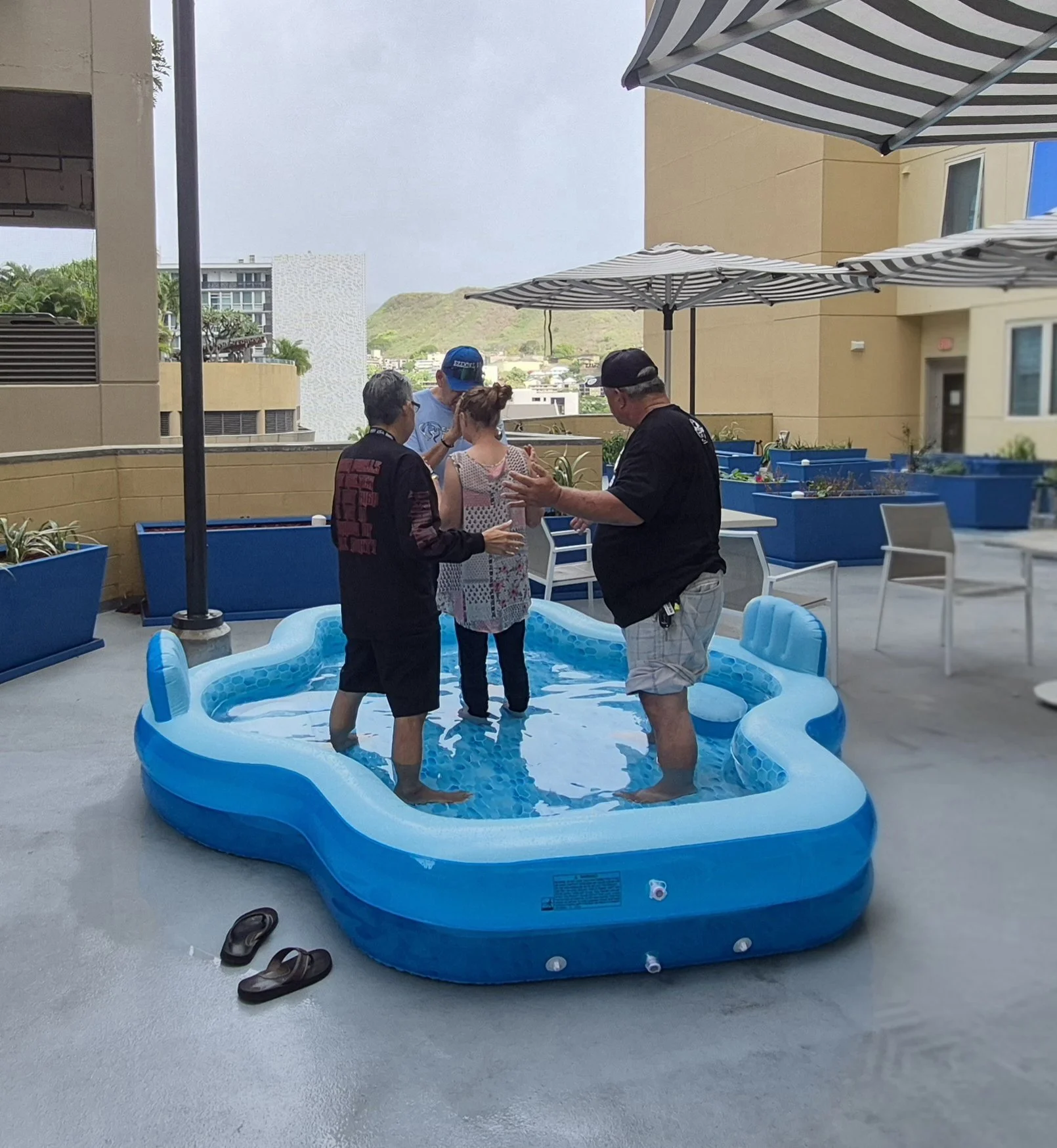 Four people standing in and around a small inflatable pool on a rooftop patio, with two women and two men, all engaged in conversation, with umbrellas and city buildings in the background.