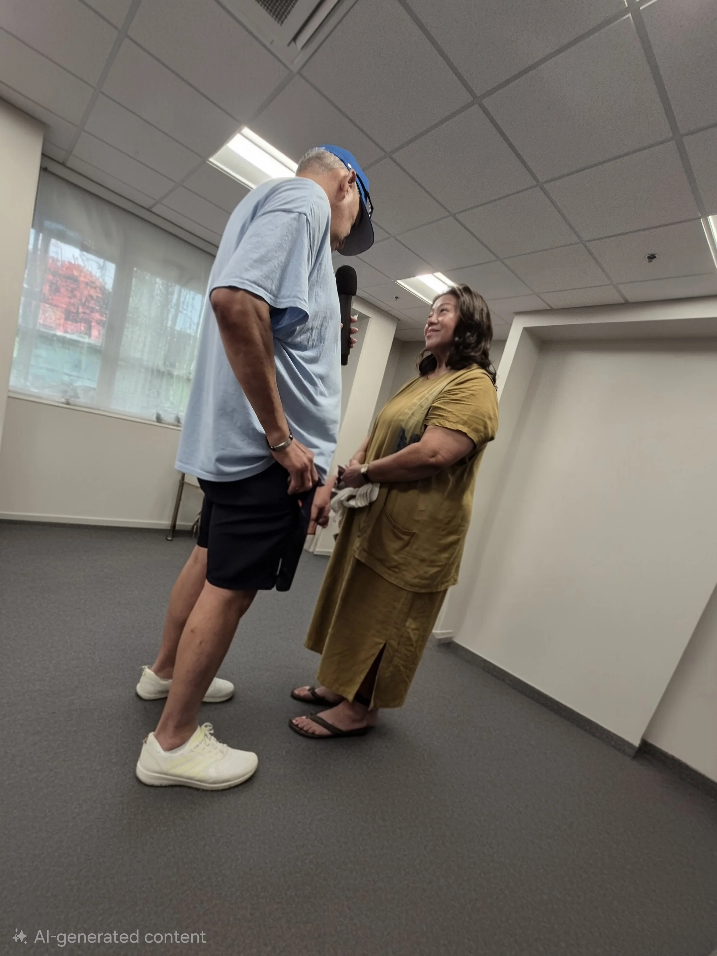 A man and woman are standing face-to-face indoors, dressed casually, engaging in a conversation. The man is holding a microphone and is slightly taller than the woman. The room has a window with a view of trees outside, a drop ceiling with fluorescent lights, and plain walls.