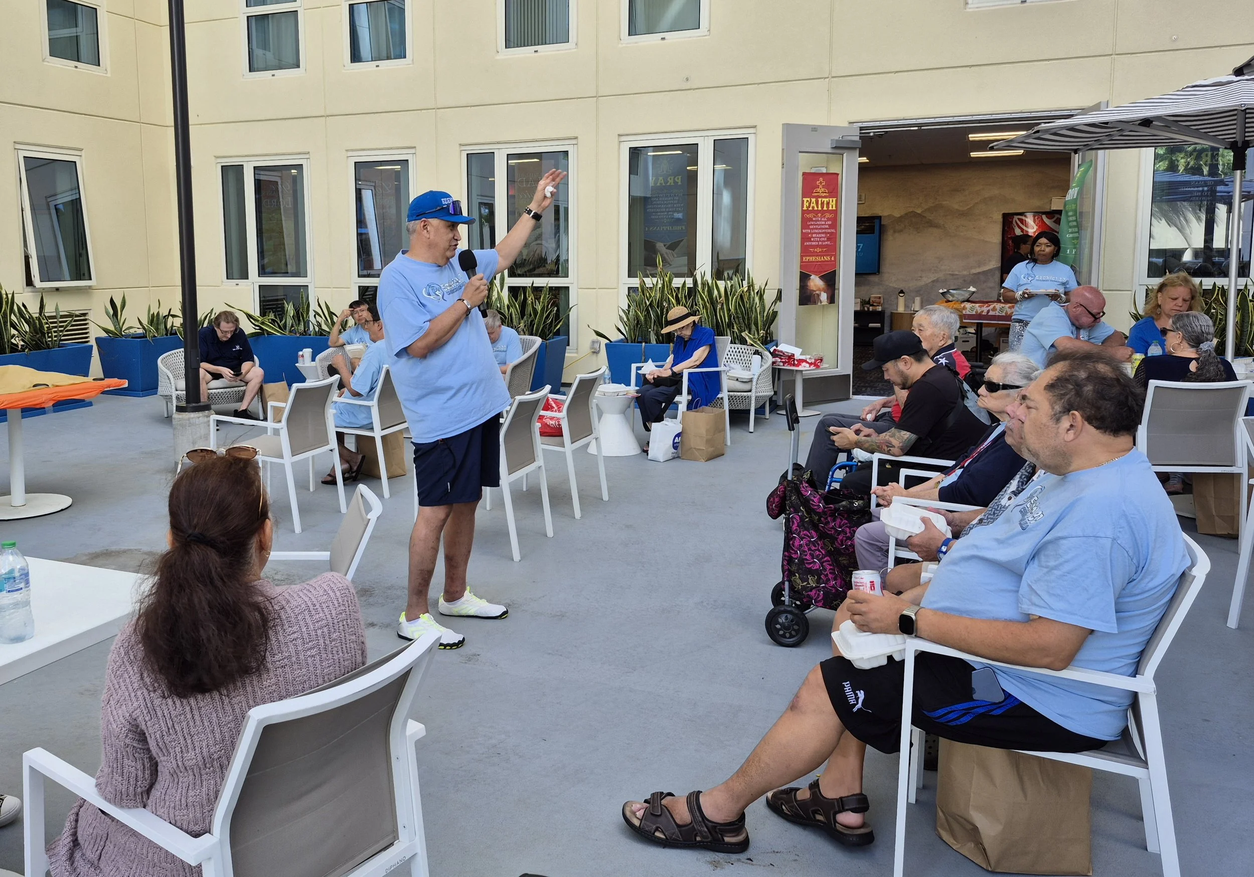 A man in a blue t-shirt, dark shorts, and a blue cap is speaking into a microphone to a seated audience on an outdoor patio. The audience includes diverse people, some with food containers, seated on chairs and benches.