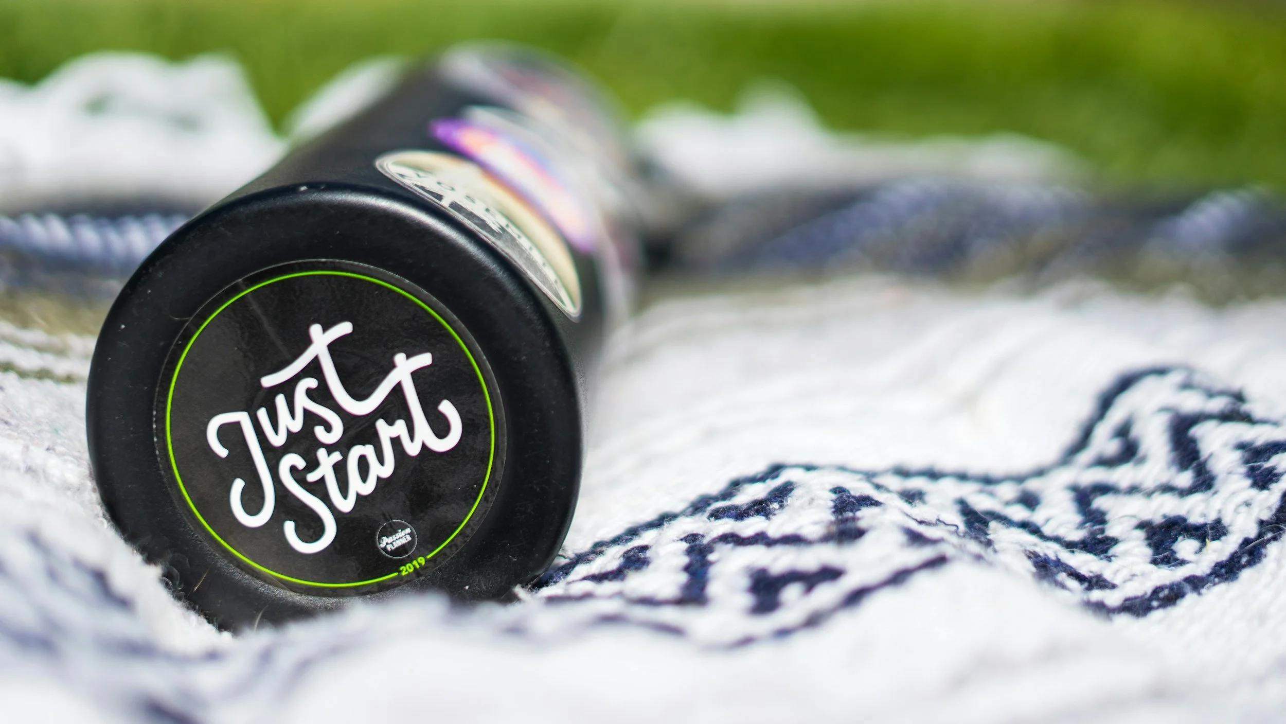 A black bottle with the words "Just Start" in white, lying on a patterned towel outdoors.