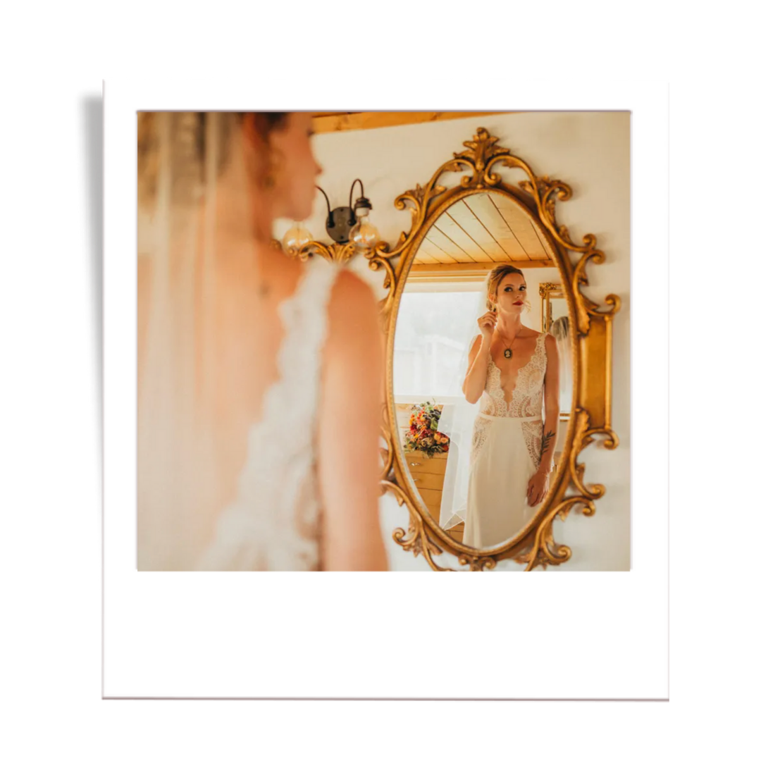 A woman in a wedding dress looking at herself in a large ornate gold-framed mirror, adjusting her earring, with a bouquet of flowers visible in the background.
