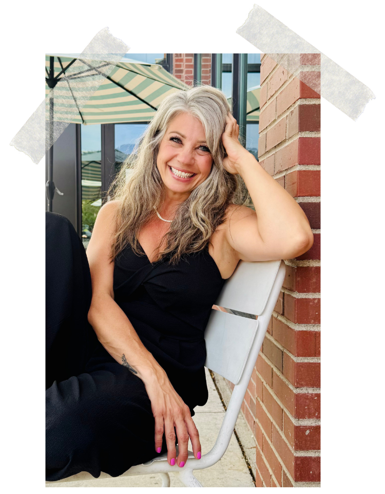 A smiling woman with long gray hair sits outside on a white chair, wearing a black dress, with one hand on her head and the other resting on her knee, next to a brick wall.
