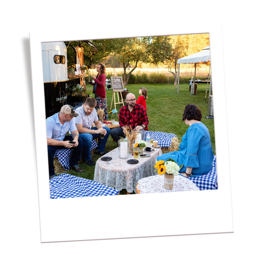 People enjoying a picnic outdoors in a grassy field with trees and tents in the background, sitting at tables with checkered tablecloths and floral decorations.