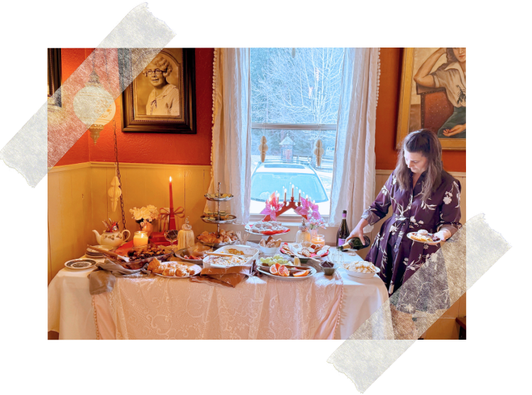 A woman at a decorated table with various dishes, cakes, and drinks, in a cozy home with orange walls, framed pictures, lace curtains, and a snowy outdoor scene visible through the window.