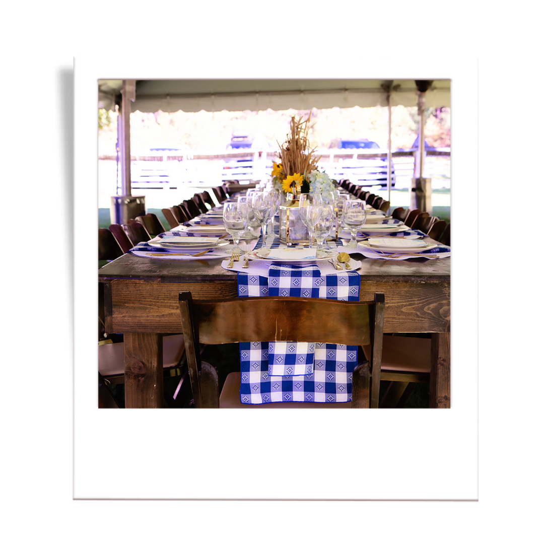 Long wooden table set for a formal dinner under a tent with a blue and white checkered table runner, floral centerpiece, and glassware.