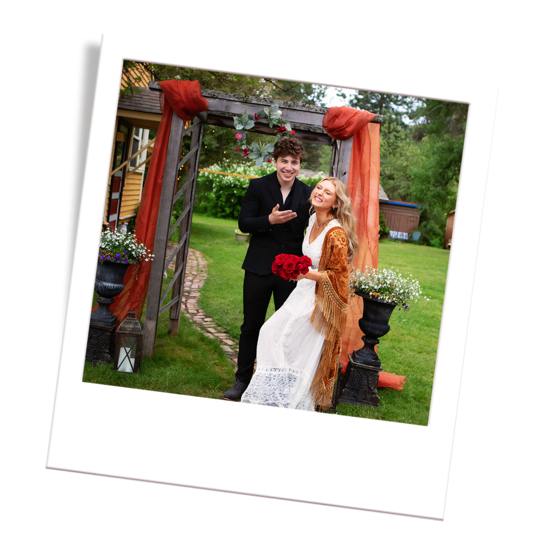A young couple at a wedding, standing under a wooden arch decorated with orange fabric and flowers, outside in a garden.