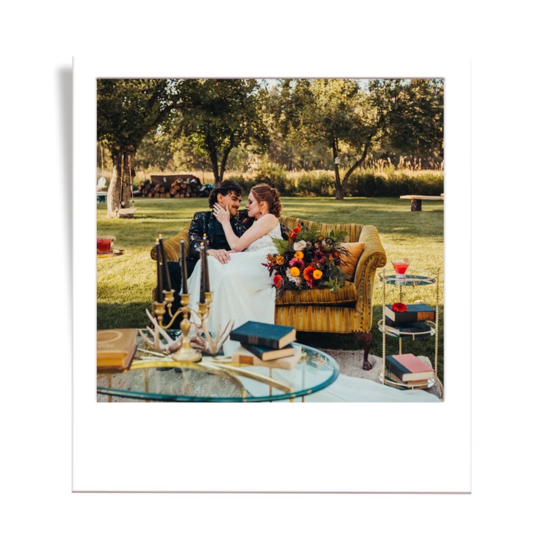 A couple sitting on a vintage yellow couch outdoors surrounded by trees, with a wedding dress and bouquet of flowers, and decorative books and candles on a glass table nearby.