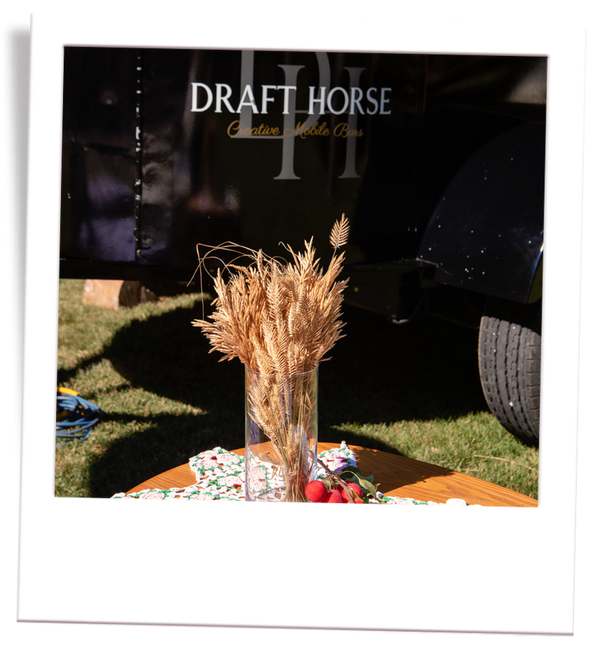 A vase with dried wheat and grass on a table, with cherries and a cloth, outside near a trailer reading 'Draft Horse Creative Mobile Bar' in the background.
