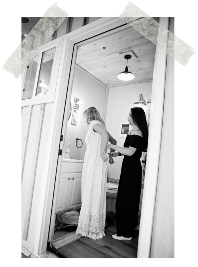 Two women in a small bathroom, one in a long white dress, the other in a black dress, with the woman in black helping the woman in white