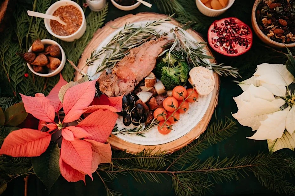 A holiday feast with a roasted meat, cherry tomatoes, broccoli, bread, and herbs on a decorative plate surrounded by poinsettias, nuts, and small bowls of sauce or relish on a green tablecloth.