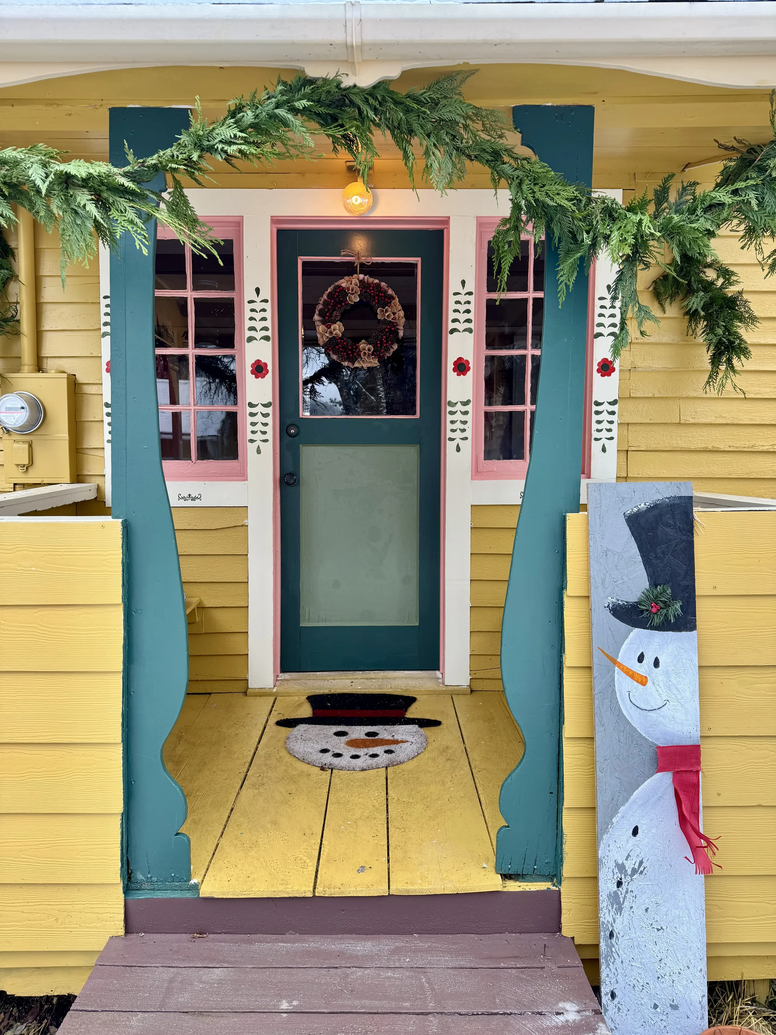 Front view of a yellow house with red trim, featuring two windows with pink frames and decorative flower patterns, a red door with a glass panel and a pink bottom panel, and red curtains on the sides.
