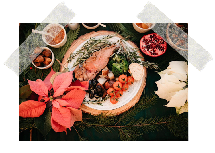 A holiday feast with a roasted meat, cherry tomatoes, broccoli, bread, and herbs on a decorative plate surrounded by poinsettias, nuts, and small bowls of sauce or relish on a green tablecloth.