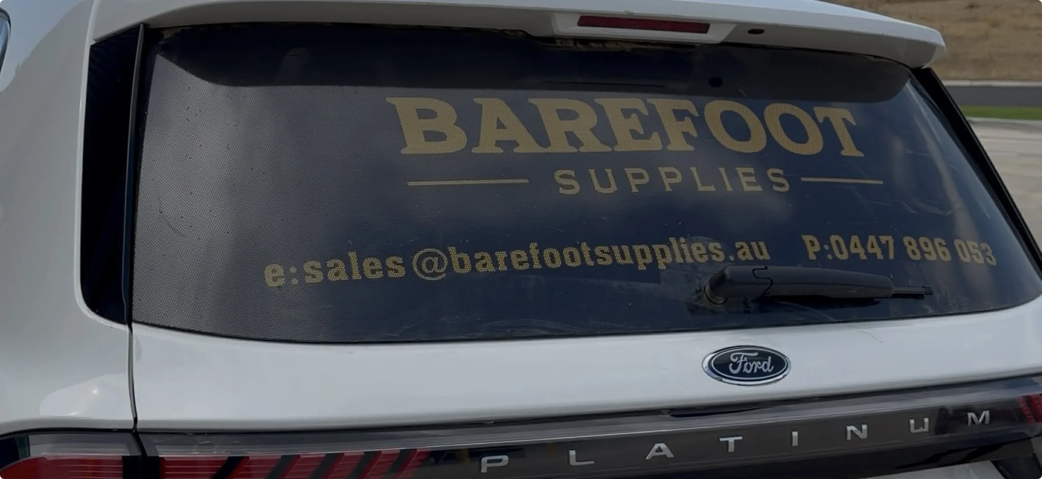 Rearview of a white Ford vehicle with a large advertisement decal on the rear window for Barefoot Supplies, including an email address and phone number.