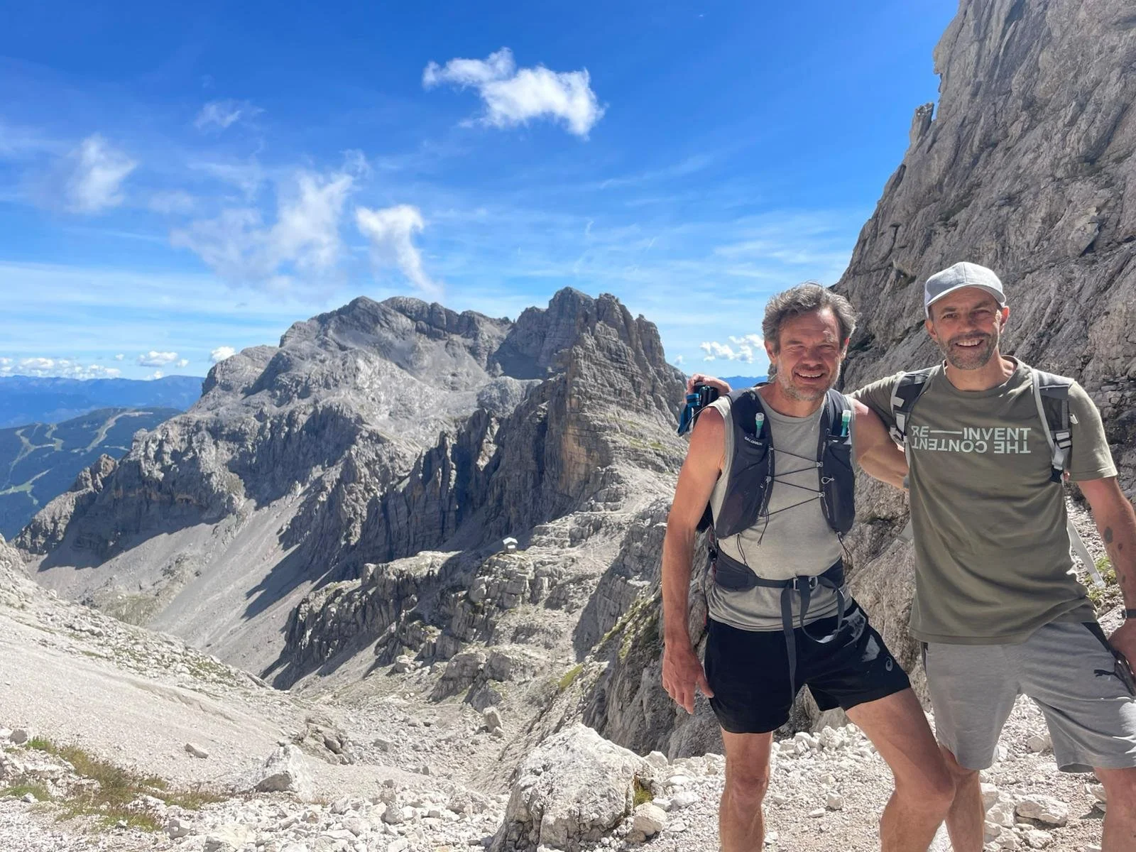 Two men smiling, posing on rocky mountain terrain with rugged peaks and a blue sky in the background, wearing hiking gear and backpacks.