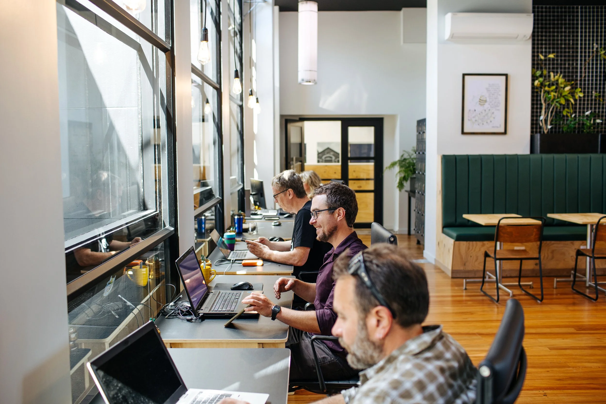 Coworking desks at The Business Hive with people sitting at desks in front of large windows, in a bright space with a wooden floor, some wooden tables and chairs and a double glass door in the background
