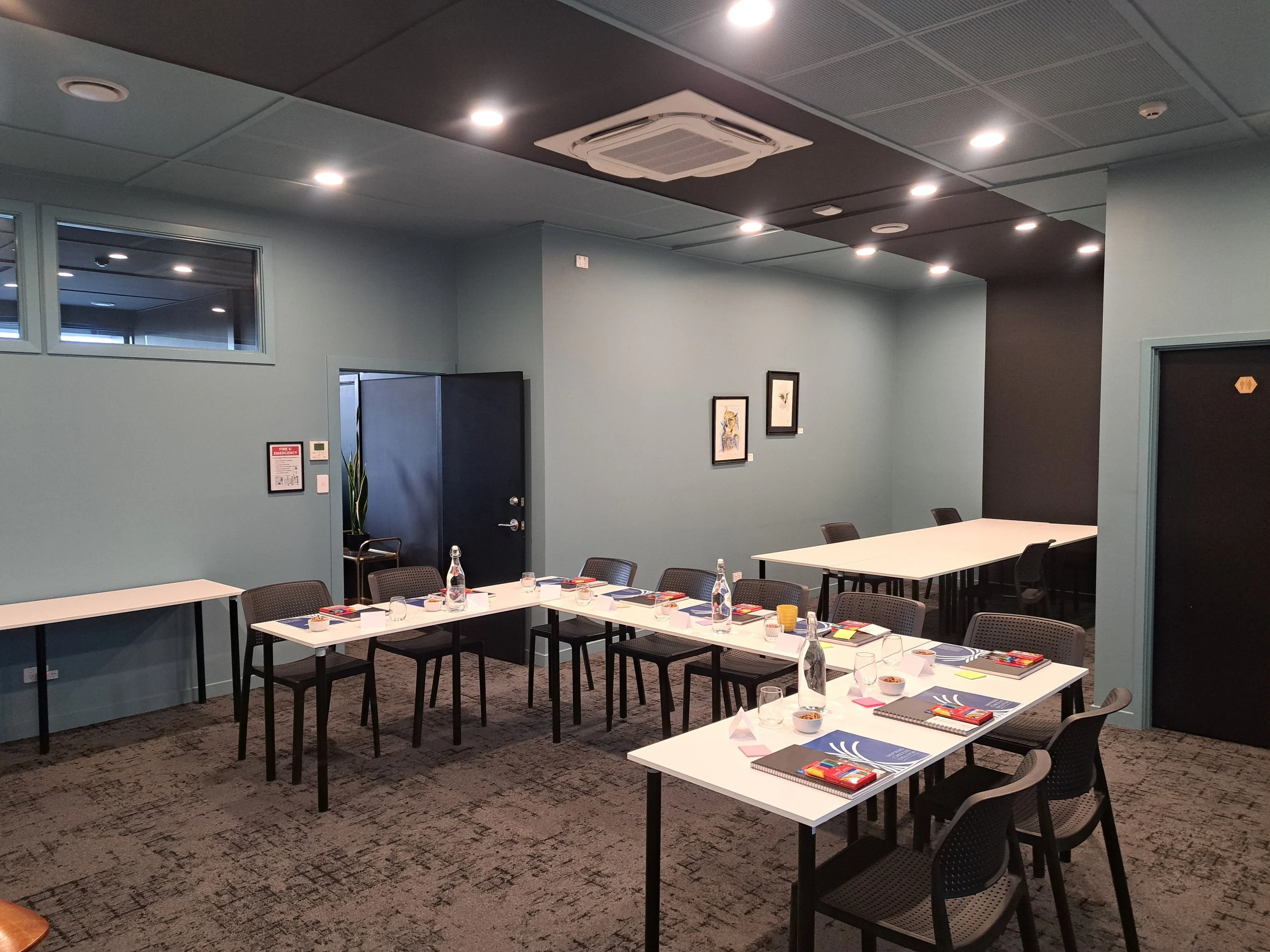 Training and meeting room with tables arranged in a U-shape, set with notebooks, pens, glasses, water bottles, bowls, and small notebook pads, ready for a presentation or workshop.
